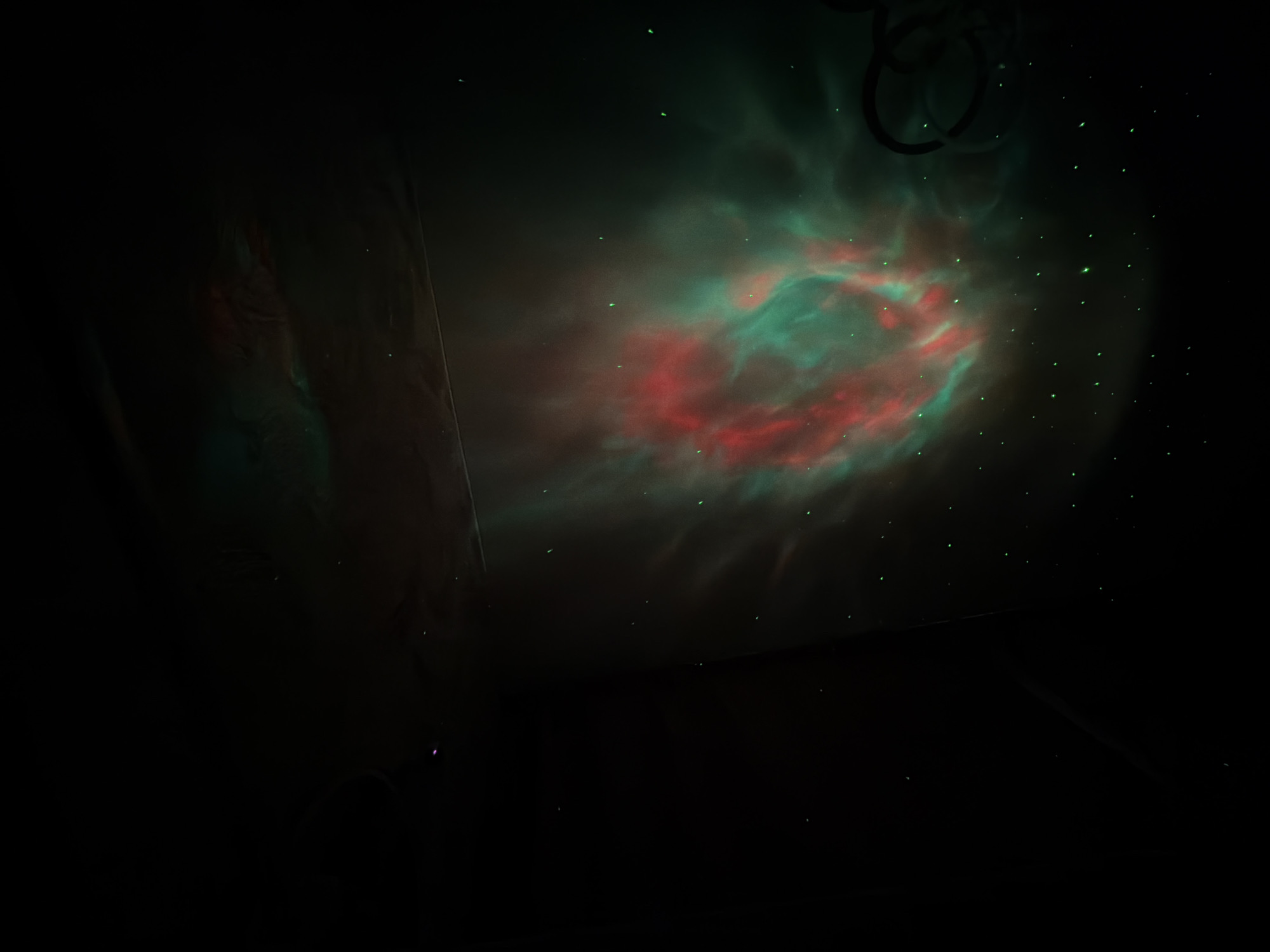 The image displays a dark indoor setting, possibly a room, illuminated by a celestial-themed light projection on a wall or ceiling. The main feature is a swirling pattern of red and green light, resembling a nebula or galaxy, with numerous small, bright green dots scattered across it, mimicking stars. The background is predominantly dark, creating a deep space effect.
In the upper right section of the projection, some dark, circular or looped structures are faintly visible, which could be part of a light fixture, hanging wires, or objects in the room. A subtle hint of what might be a dark curtain or fabric edge can be seen on the left side of the frame, further indicating an enclosed space. The overall impression is one of an atmospheric and decorative light display, likely used for ambiance or relaxation. There are no people or explicit activities depicted, and no visible text. The time of day is inferred to be dark, allowing the projection to be clearly visible. The specific location of Reshetnikovo, Russia, is provided as context but is not visually identifiable within the image itself.