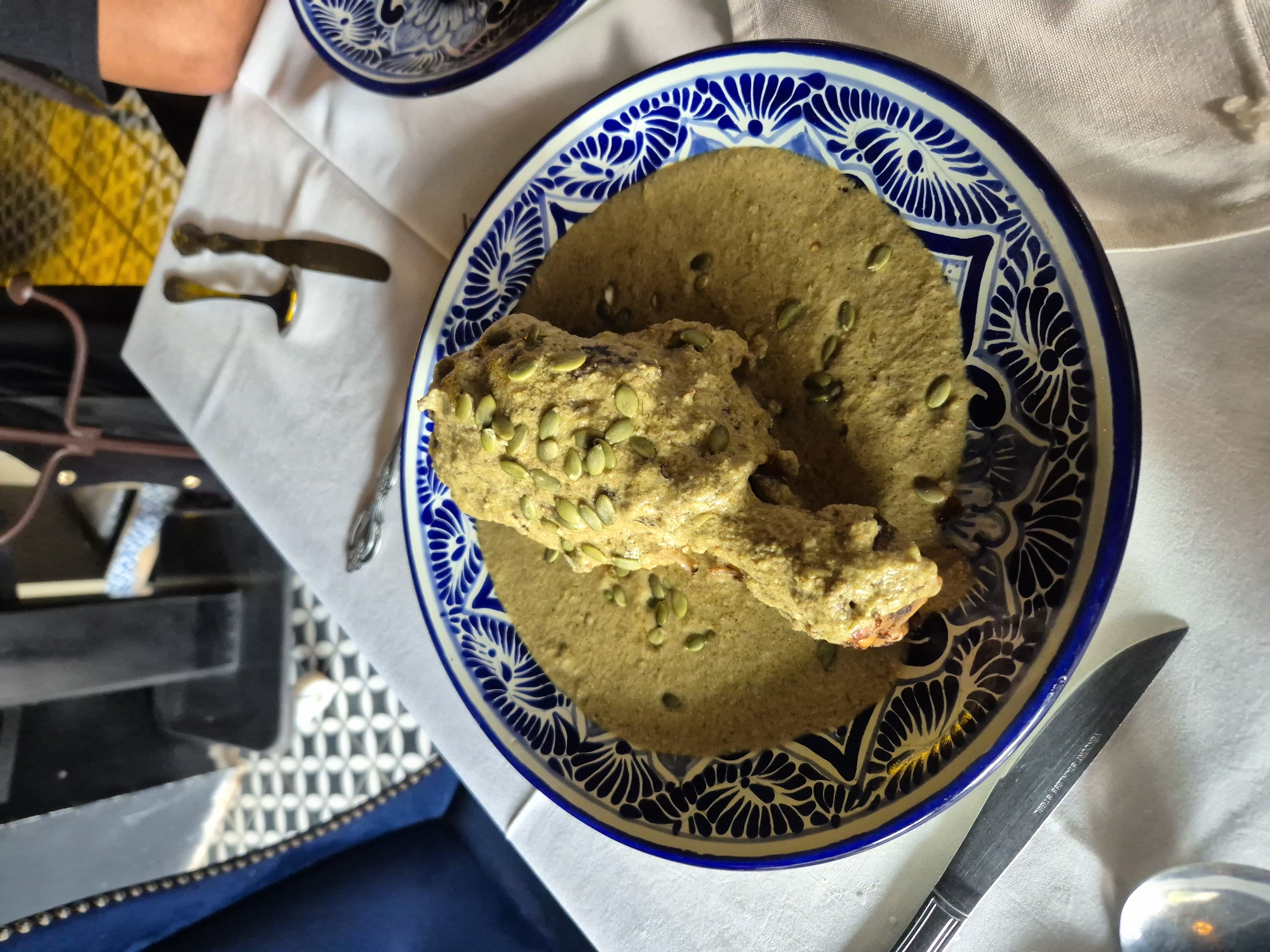 The image shows a plate of Mole Poblano, a traditional Mexican dish, likely photographed in Puebla, Mexico. The dish is presented on a blue and white Talavera-style ceramic plate, with a chicken drumstick covered in mole sauce, topped with pumpkin seeds. The plate sits on a white tablecloth. A butter knife and spoon are located next to the plate, along with the edge of a blue chair with silver studs. Other background elements include patterned walls and dining furniture.