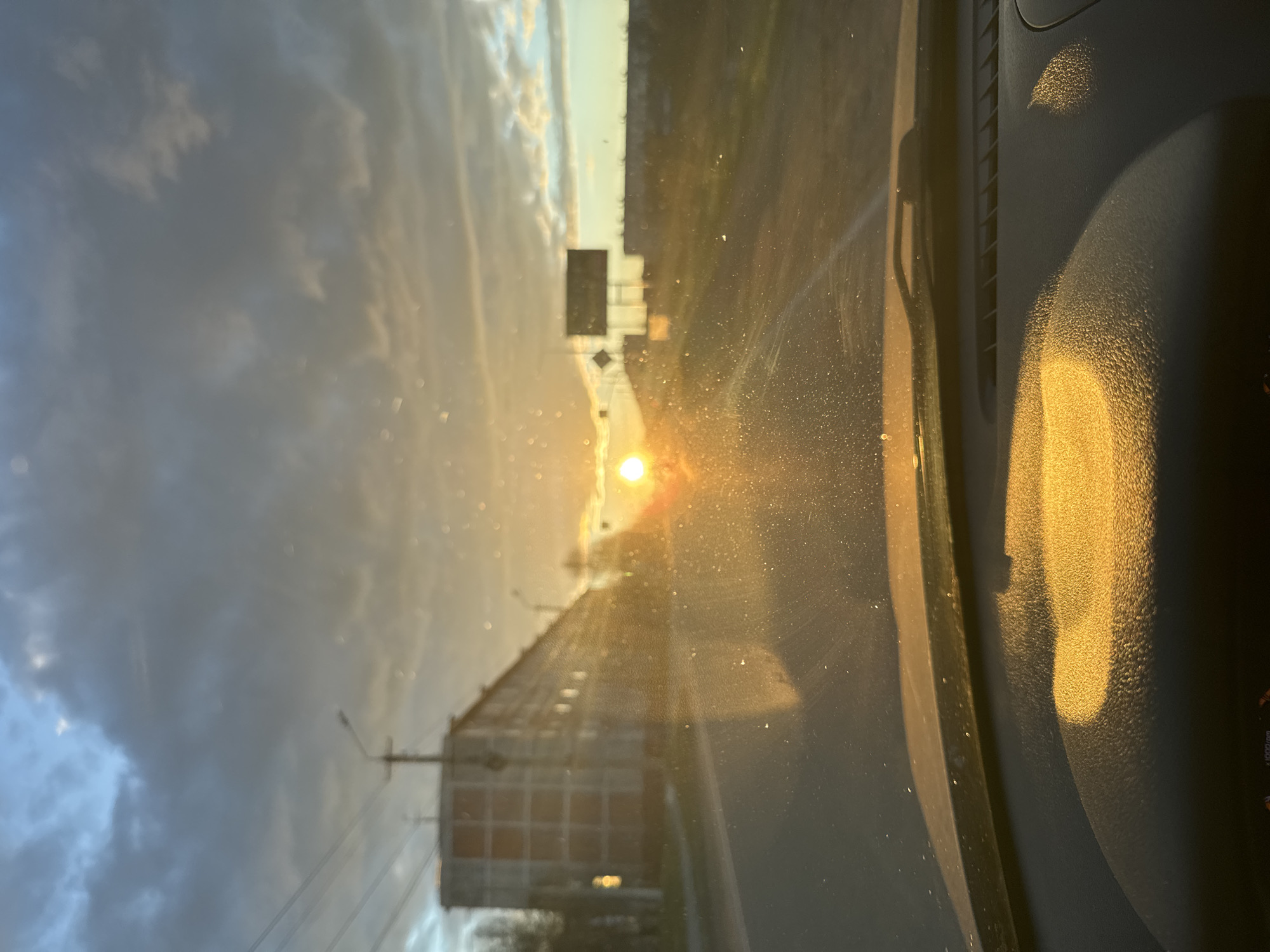 The image, taken from inside a vehicle, depicts a sunset along a road in Apatity, Russia. The setting sun is centered in the windshield's view, creating glare and obscuring details. To the left, a multi-story building is visible. Streetlights and overhead wires are also present. Road signs are partially visible in the distance, likely indicating traffic directions. The car's dashboard and interior elements are visible in the foreground. The general environment is urban, with a mixture of residential and roadside infrastructure.