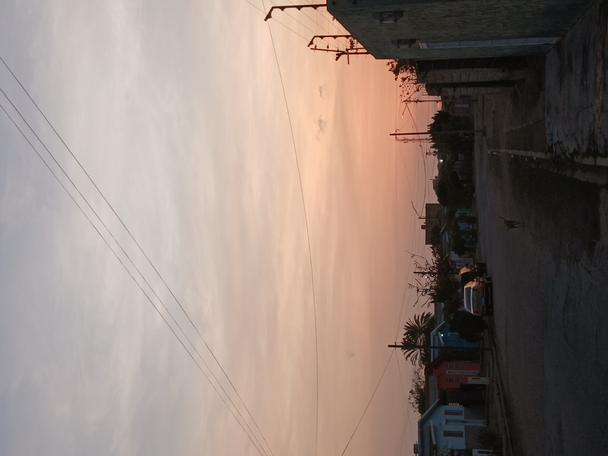 The image shows a street in Juan Griego, Venezuela at dusk. The sky is a pale orange and grey. Utility wires crisscross the sky. The street is paved and runs between low buildings. Some of the buildings are brightly colored. Cars are parked on the side of the road.
