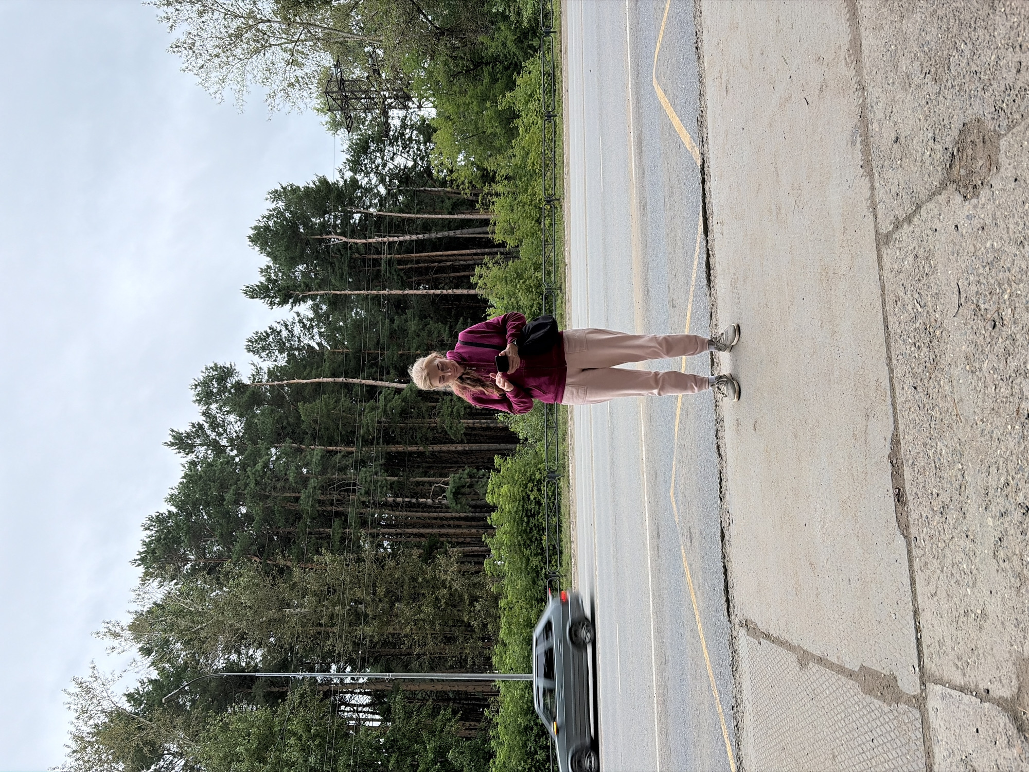 A woman stands on a concrete sidewalk at a bus stop next to a road. She is wearing a maroon hooded sweatshirt, pink sweatpants, and grey sneakers. She is holding a smartphone and looking at it. A black bag is slung over her right shoulder. In the background, there is a line of pine trees, a street light, and a power line tower. A grey car is driving past on the road, blurred due to motion. The scene appears to be a rural or suburban area on an overcast day. This location is likely the bus stop on Votkinskoye Highway in Chaikovsky, Russia, given the characteristic pine forest scenery common in the region.