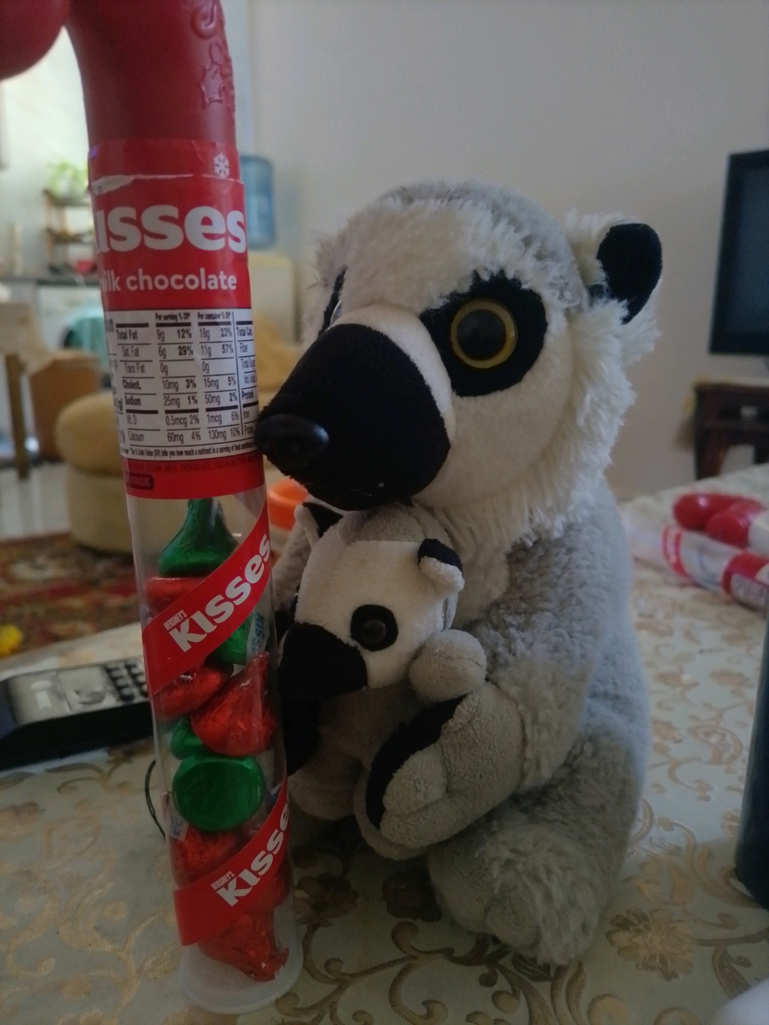 A stuffed lemur toy sits on a patterned tablecloth next to a tube of Hershey's Kisses. The lemur is gray and white with large yellow eyes and black accents. It is holding a smaller stuffed animal, possibly a baby lemur, in its arms. The tube of Hershey's Kisses is red and clear, showing red and green foil-wrapped candies inside. The background is blurred but appears to be an indoor setting with furniture and a television. The image is taken from a close-up perspective, focusing on the toys and the candy. There is no indication of Amman, Jordan, or any specific city.