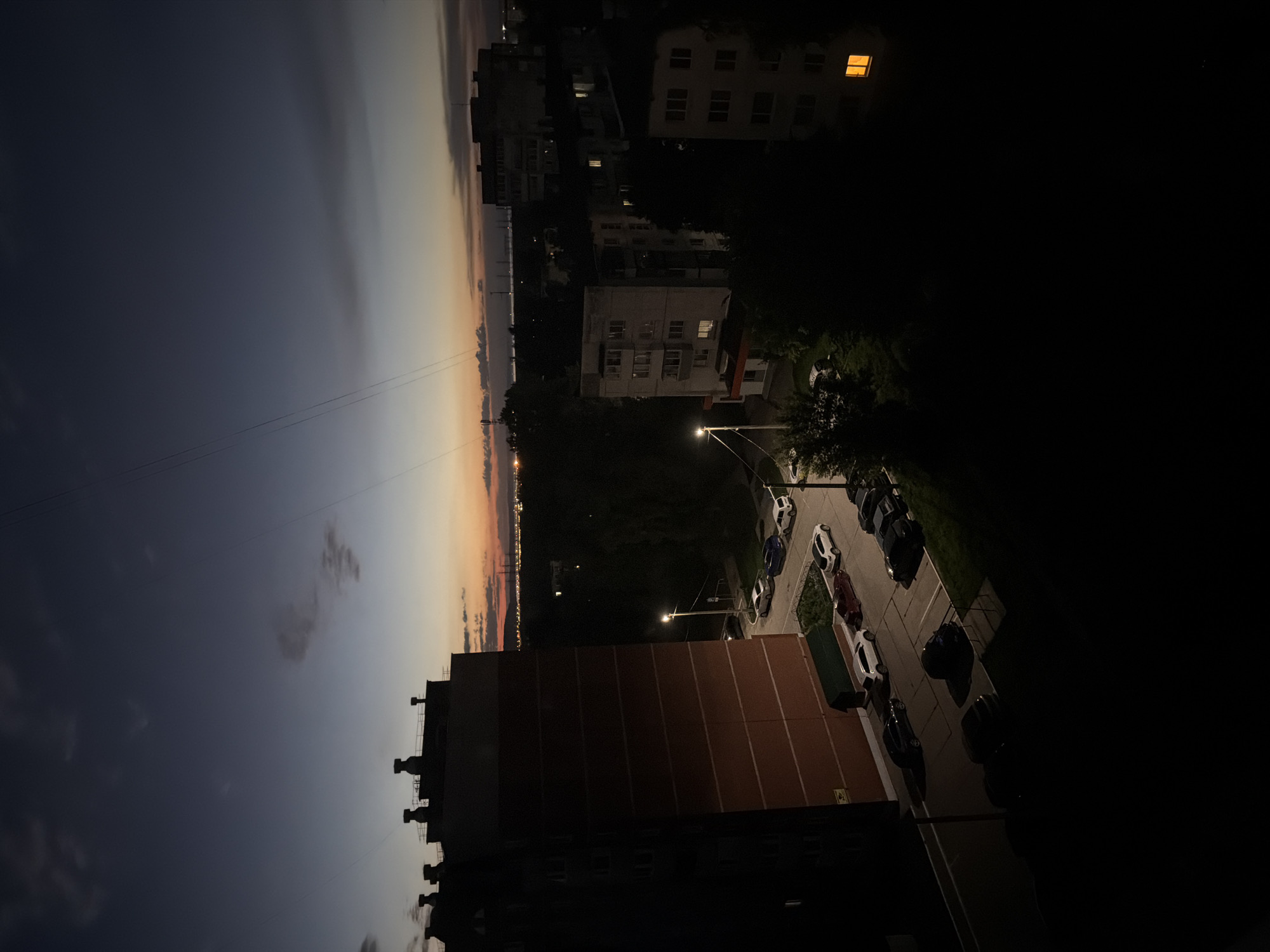 The image captures a twilight scene in the city of Chaikovsky, Russia, as viewed from a high vantage point, likely a residential building. The sky displays a gradient of deep blue at the top, transitioning to lighter blues and oranges near the horizon, indicating dusk. Several apartment buildings are visible, with the most prominent being a tall, brick-clad building on the left, partially in shadow.
Below the buildings, a street and parking area are visible, with several cars parked. These include white, dark blue, and black sedans, along with a white SUV. Streetlights illuminate parts of the area, casting long shadows.
In the background, more apartment buildings and trees can be seen. The distant horizon features a faint glow of city lights and industrial structures, suggesting the presence of a larger urban or industrial area. The overall atmosphere is quiet and still, characteristic of a residential neighborhood at the end of the day. There are no people visible in the image.