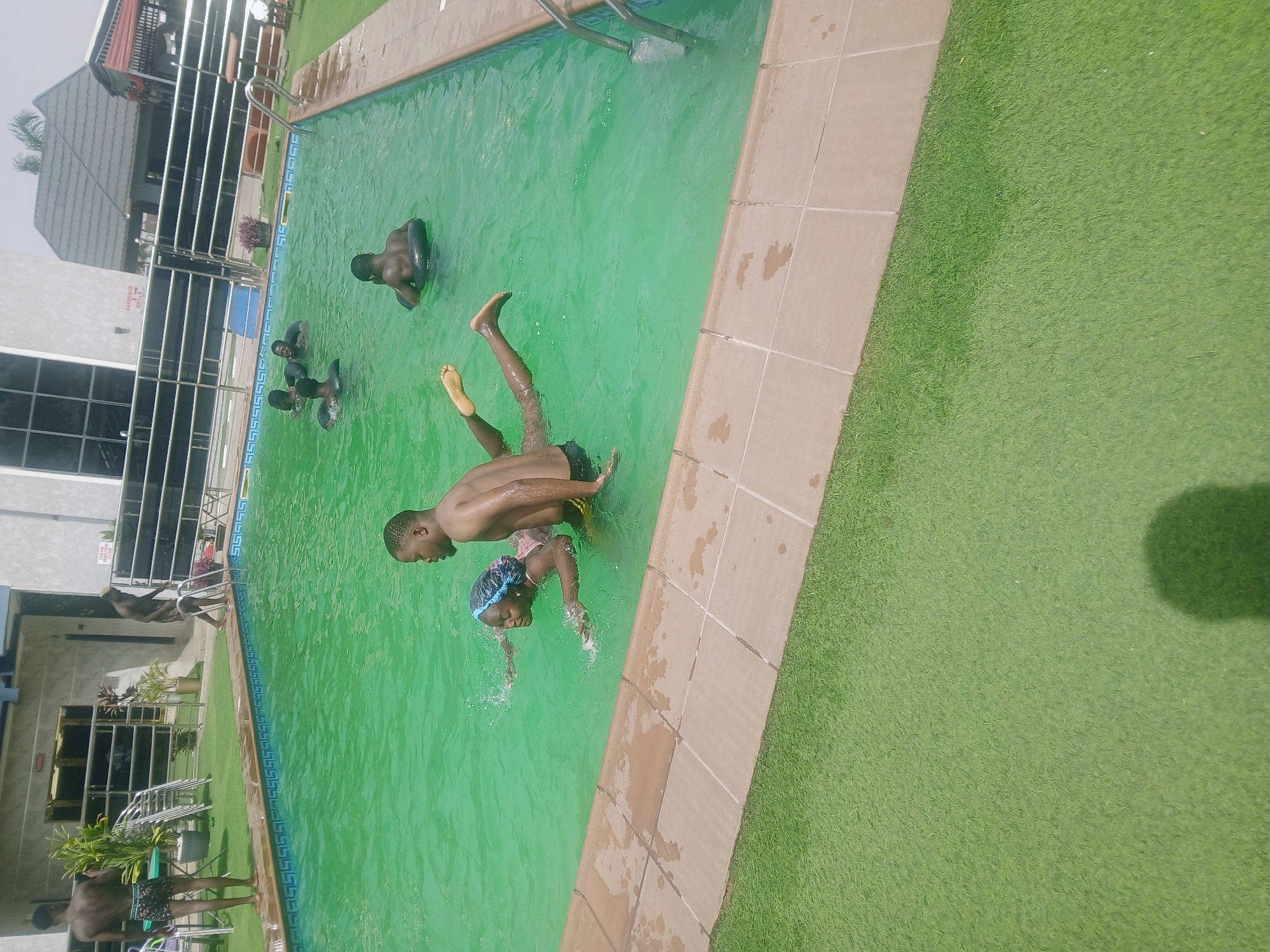The image shows a group of people swimming in a green-colored swimming pool, seemingly located outdoors in Bukuru, Nigeria. Two people are interacting at the edge of the pool, one appearing to be helping the other into the water. Others are in the pool, some with inner tubes. A building is visible in the background. The immediate surrounding area of the pool has tile and artificial turf.