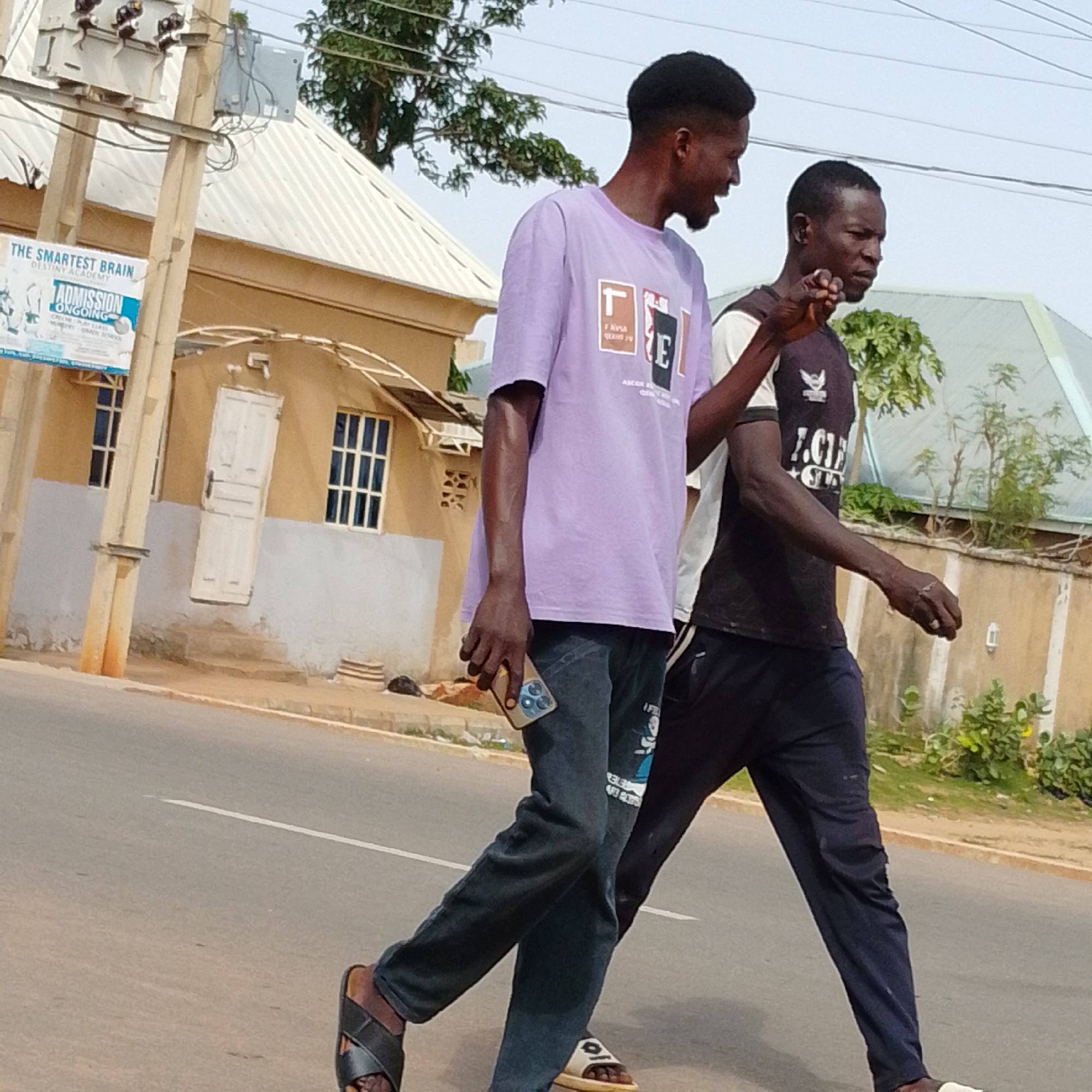 The image depicts two young men walking on a paved road in Gombe, Nigeria. The man on the left, wearing a light purple t-shirt and dark pants, is holding a smartphone in his right hand. The man on the right, wearing a dark t-shirt with white and black accents and dark pants, is gesturing with his right hand. They are walking side-by-side, with the man on the left slightly ahead. In the background, there is a light brown building with a white door and windows, and a signpost on the left side of the image that reads "THE SMARTEST BRAIN DESTINY ACADEMY ADMISSION ONGOING". Trees and vegetation are visible behind the building and along the roadside. The sky is overcast. The overall scene appears to be a typical daytime street view in a residential or commercial area.