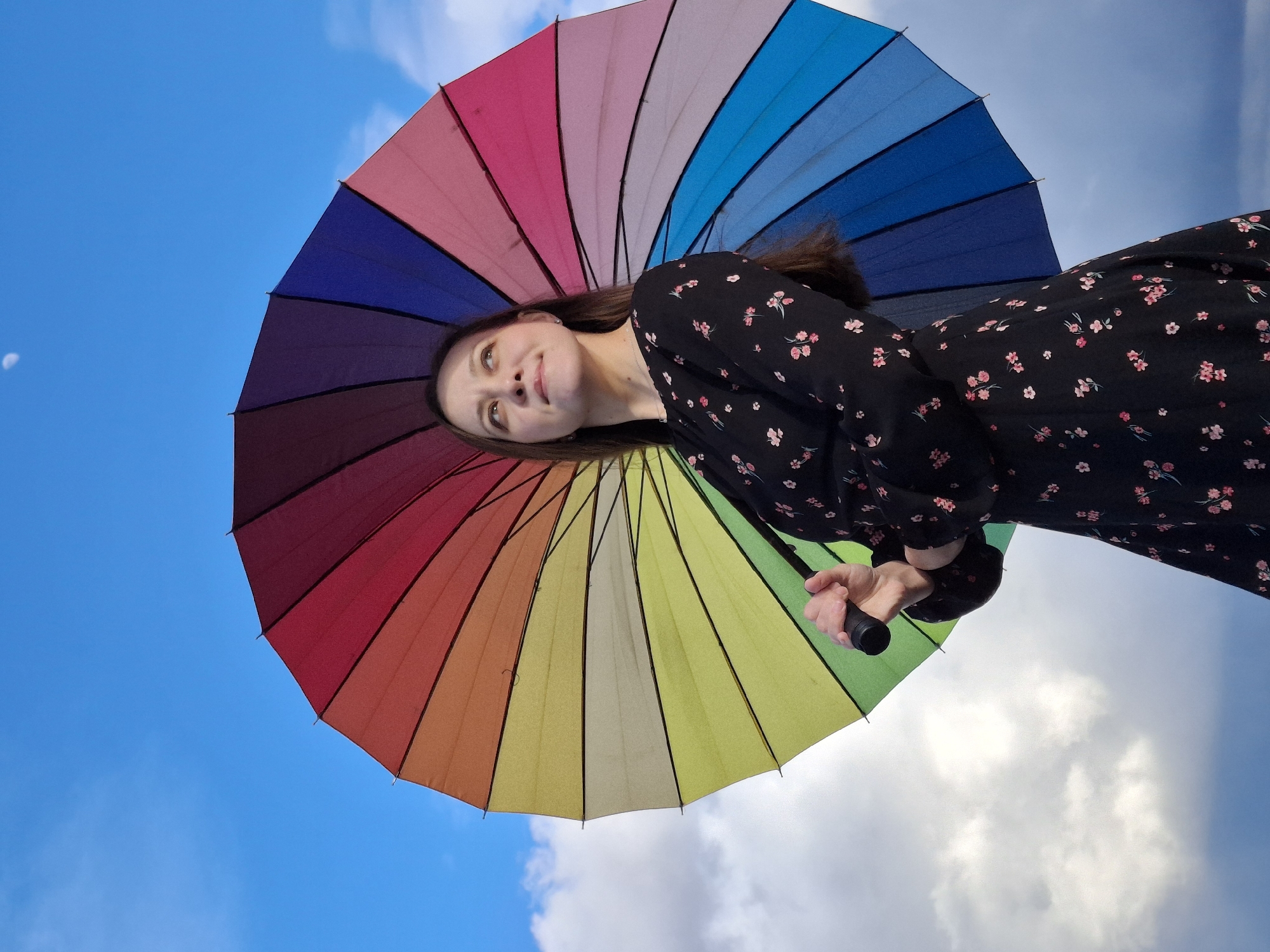 A low-angle, portrait-oriented shot depicts a woman holding a multi-colored umbrella against a bright blue sky with some white clouds. The umbrella's color segments progress from red, orange, yellow, green, and blue. The woman has brown hair and is wearing a dark-colored dress with a small pink floral pattern. She's looking up towards the top left corner of the frame. A faint moon appears in the upper left portion of the sky. The image gives no explicit information to suggest Chaikovsky, Russia as the location.