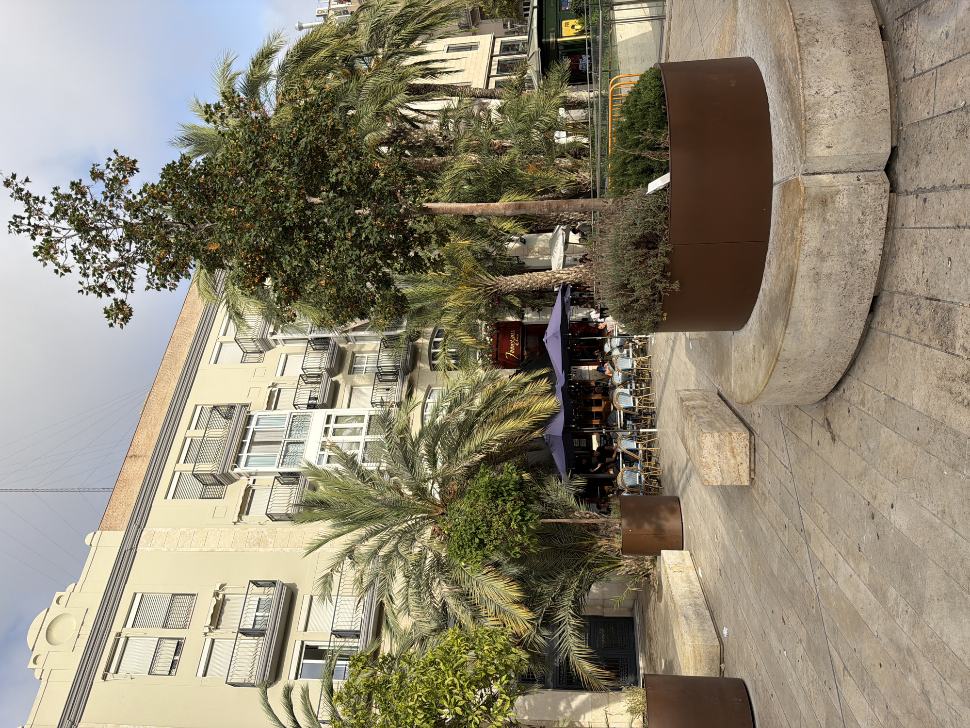 An outdoor cafe area is set up in front of a pale yellow building with multiple balconies. Several palm trees and a tall tree with orange-colored fruit are visible. Tables and chairs are arranged on the pavement, with some customers seated at the tables. A large, curved concrete bench with a brown planter box containing greenery surrounds the tall tree. The ground is paved with light-colored stone tiles.