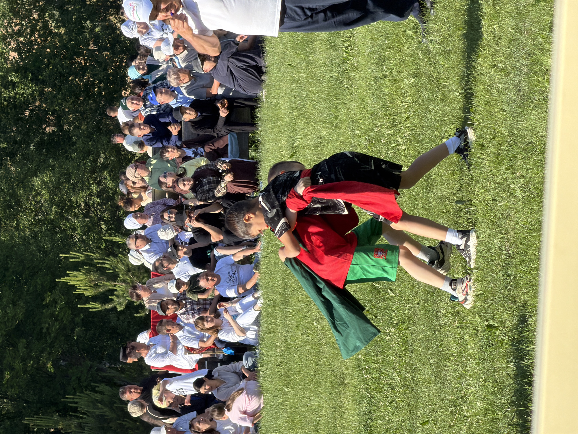 This media file captures an outdoor event under what appears to be bright daylight. The scene features a gathering of people, predominantly adults and children, spread across a grassy area. In the foreground, two children are actively engaged in what looks like a game or a friendly wrestle on the green grass. One child is wearing a red shirt and green shorts, while the other is in a black shirt. Both are wearing athletic shoes with white socks. Their bodies are positioned in a way that suggests movement and interaction.
Behind the children, a large crowd of spectators is visible. They are seated or standing on the grass, facing towards the activity in the foreground. The crowd is diverse in age and attire, with some individuals wearing hats or head coverings. The expressions on their faces are generally attentive, suggesting they are watching the event unfold.
To the left of the crowd, dense green foliage, likely trees and bushes, forms a natural backdrop, indicating an outdoor setting such as a park or a field. The light suggests it's daytime, possibly morning or afternoon, given the clear skies and visible shadows. There are no prominent objects or texts that provide specific location cues or brand information. The overall atmosphere appears casual and lively, typical of a community gathering or a sporting event.