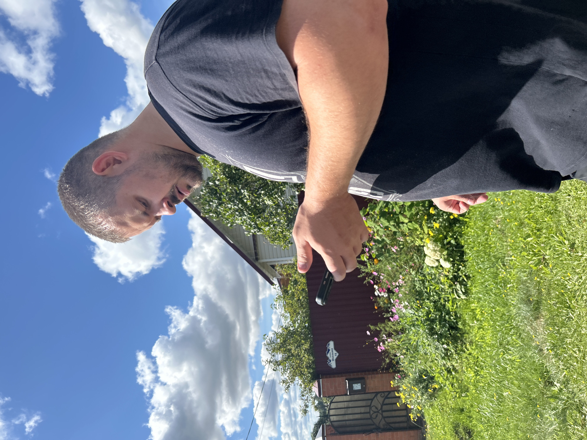 A man in a black t-shirt is standing outdoors in front of a fence with a house behind it. He is looking down at a black smartphone he is holding in his right hand. The background includes a green lawn, a flower garden, a brick and metal gate, and a building with a red roof. The sky is blue with white clouds. The location is identified as Kalitsino, Russia.