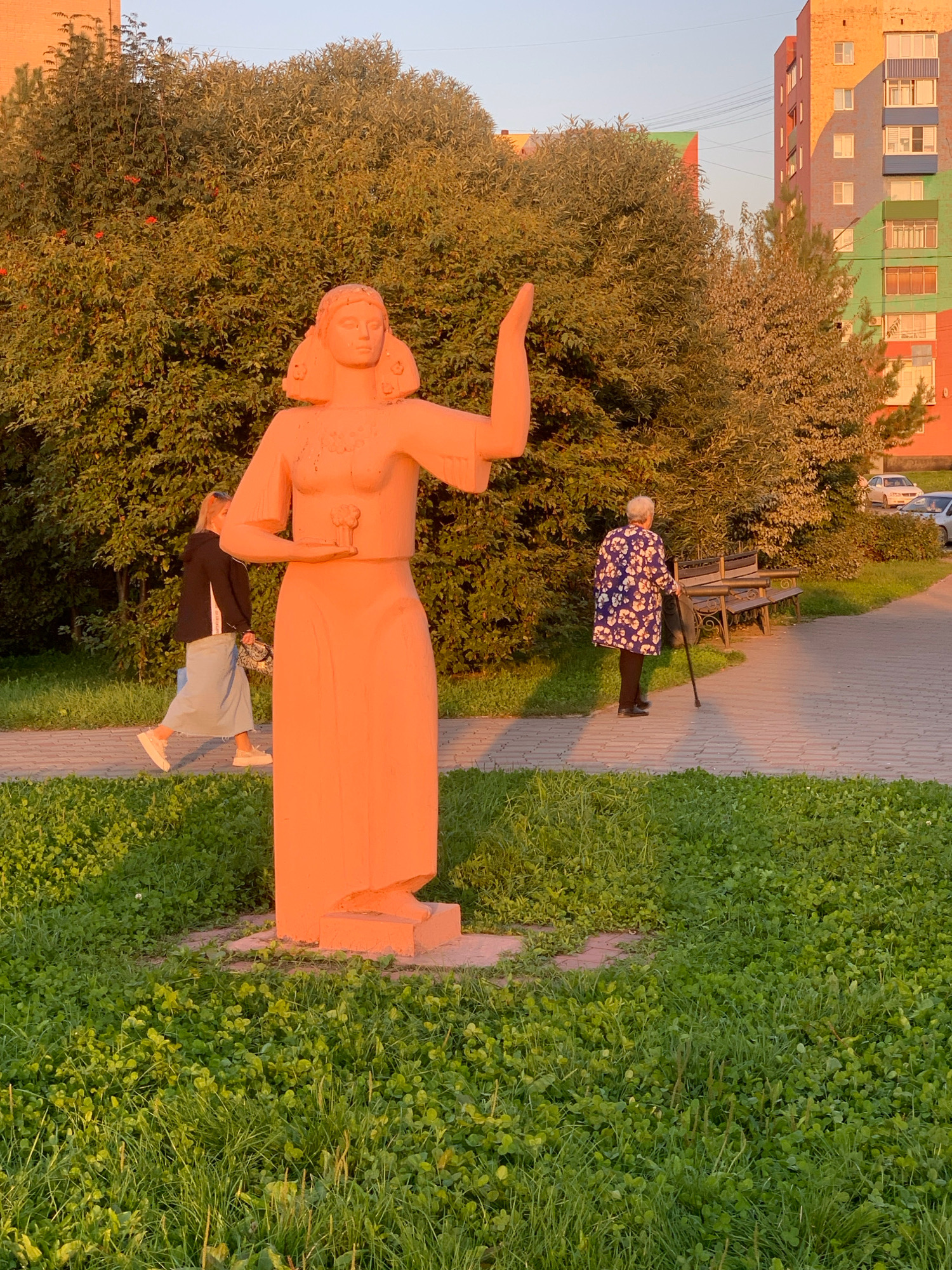A bright orange, two-dimensional statue of a woman stands in the foreground of a park-like setting. The statue depicts a woman with a round face, arms outstretched, and holding a small object in her left hand. She wears a long, flowing dress and has her hair styled around her head. Behind the statue, a paved walkway meanders through a grassy area. Two individuals are walking on the path. To the left, a woman with blonde hair, wearing a black jacket and a light blue skirt, walks away from the camera. To the right, an elderly woman with grey hair, dressed in a blue and white floral coat and black pants, walks towards the camera, leaning on a cane. In the background, a dense green hedge lines the path. Beyond the hedge, a colorful, multi-story apartment building with vibrant yellow, green, and blue sections is visible, suggesting an urban environment. The scene is illuminated by the warm glow of late afternoon or early evening sunlight. This image was taken in Prokopyevsk, Russia.