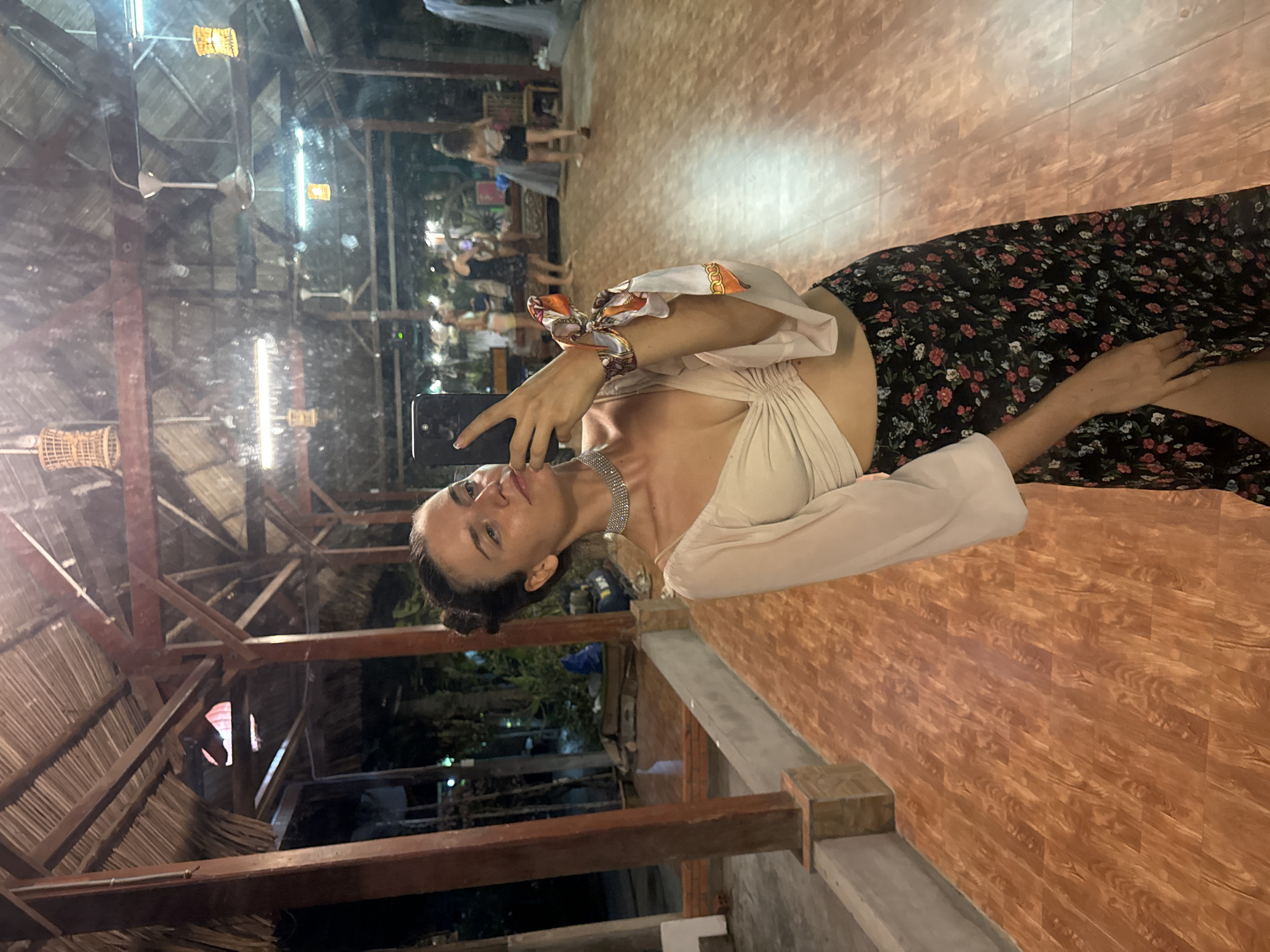 A woman takes a selfie indoors in Phu Thuy Ward, Vietnam. She wears a light-colored top, a floral skirt, and a sparkly necklace. Her hair is in a bun. She holds her phone in her left hand and looks into the screen.
The setting appears to be a restaurant or event space with a wooden roof structure and overhead lights. Other people are faintly visible in the background. The floor is a wood-look surface. There is a concrete ledge or step in the foreground.