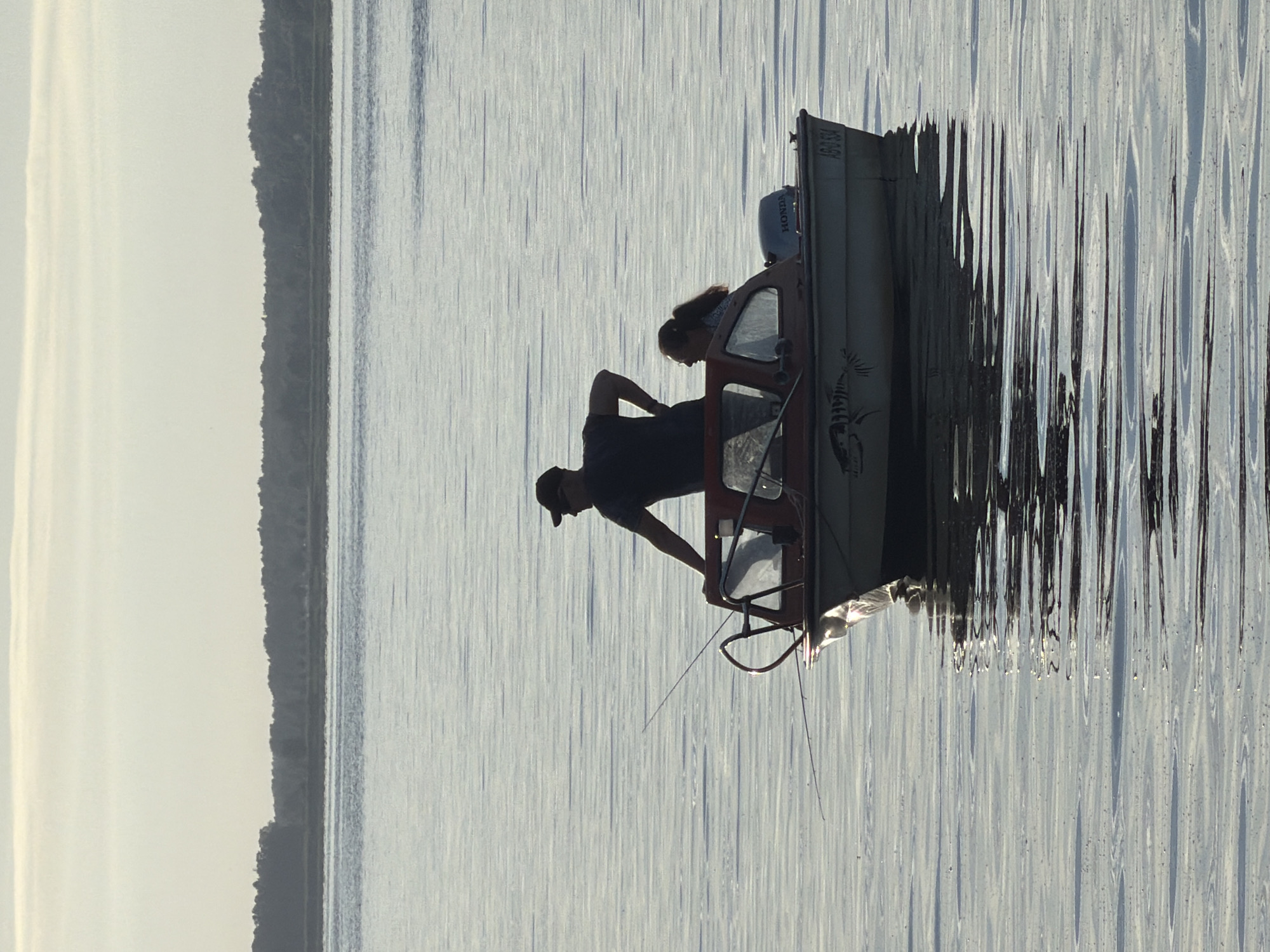 The image shows a side view of a small fishing boat on calm water. The boat is oriented vertically in the frame, suggesting the photo may have been rotated. Two people are visible in silhouette within the boat. One person, likely male, is wearing a baseball cap and appears to be leaning over something inside the boat. Another person, possibly female, with long hair pulled back, is standing beside them, looking down. The boat has a cabin with a windshield and a dark-colored interior. A Honda outboard motor is mounted on the back of the boat. The water is serene with subtle ripples, and the sky is a pale, bright color, indicating it could be early morning or late afternoon. The reflection of the boat and the surrounding environment on the water is distorted by the ripples, creating an abstract pattern. On the left side of the image, a dark, tree-lined shore is visible, suggesting a lakeside or coastal setting. The overall mood is peaceful and quiet. There is some visible text on the stern of the boat that appears to be a license plate number, reading "ABD-X531". The location context is Ribnitz-Damgarten, Germany.