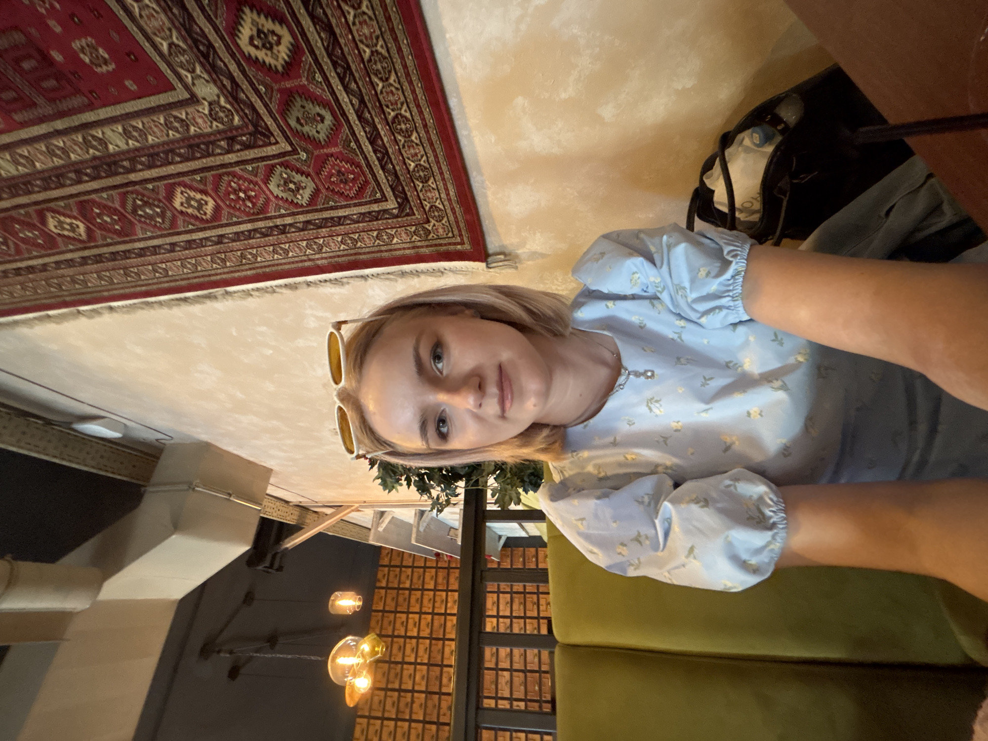 A young adult female, identified as the primary subject, is seated indoors in what appears to be a cafe or restaurant in Saint Petersburg, Russia. She possesses light blonde, shoulder-length hair with white-framed sunglasses positioned on top of her head. Her gaze is directed towards the viewer, exhibiting a neutral expression with a slight smile. She is attired in a light blue blouse featuring puffed, elasticized short sleeves and a subtle yellow floral pattern. A silver-colored necklace with a small, clear or pearl-like pendant is visible around her neck.
The subject occupies a vibrant lime-green upholstered booth seat. To her right, a black handbag containing a white garment and a light blue bottle is partially visible on the seat. A dark brown table surface, with a dark straw and clear glass, occupies the lower right foreground. Behind her right shoulder, a dark green artificial plant is situated.
The background features a textured, light yellow-orange wall on the right, adorned with a large, rectangular red and brown patterned rug of traditional design, including fringe along its left edge. To the left, behind a dark wooden railing, a wall with numerous small, dark wooden rectangular compartments or panels is visible. The overhead space reveals a dark ceiling from which industrial ventilation ducts extend, accompanied by several hanging light fixtures emitting a warm glow from exposed filament bulbs. The overall lighting is warm and artificial.