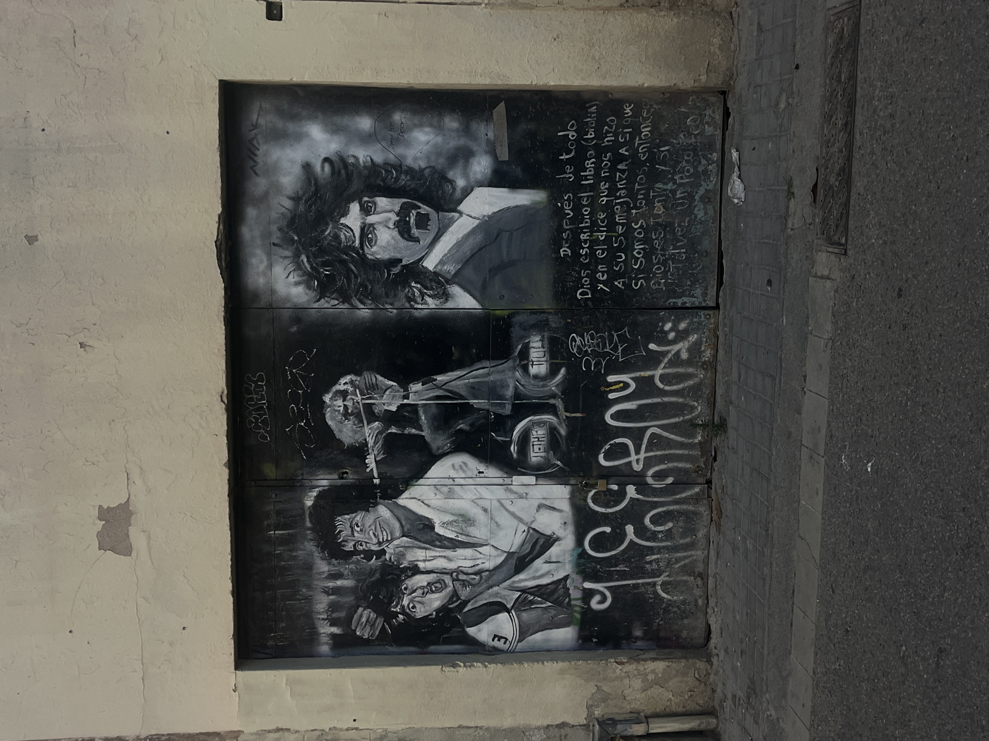 The image shows a metal door in Barcelona, Spain. The door is painted with a black and white mural featuring a portrait of Frank Zappa above figures resembling football players. Graffiti is scrawled over the artwork. To the left of the door is a wall with peeling paint, and to the right is a stone-tiled surface, presumably a sidewalk or street.