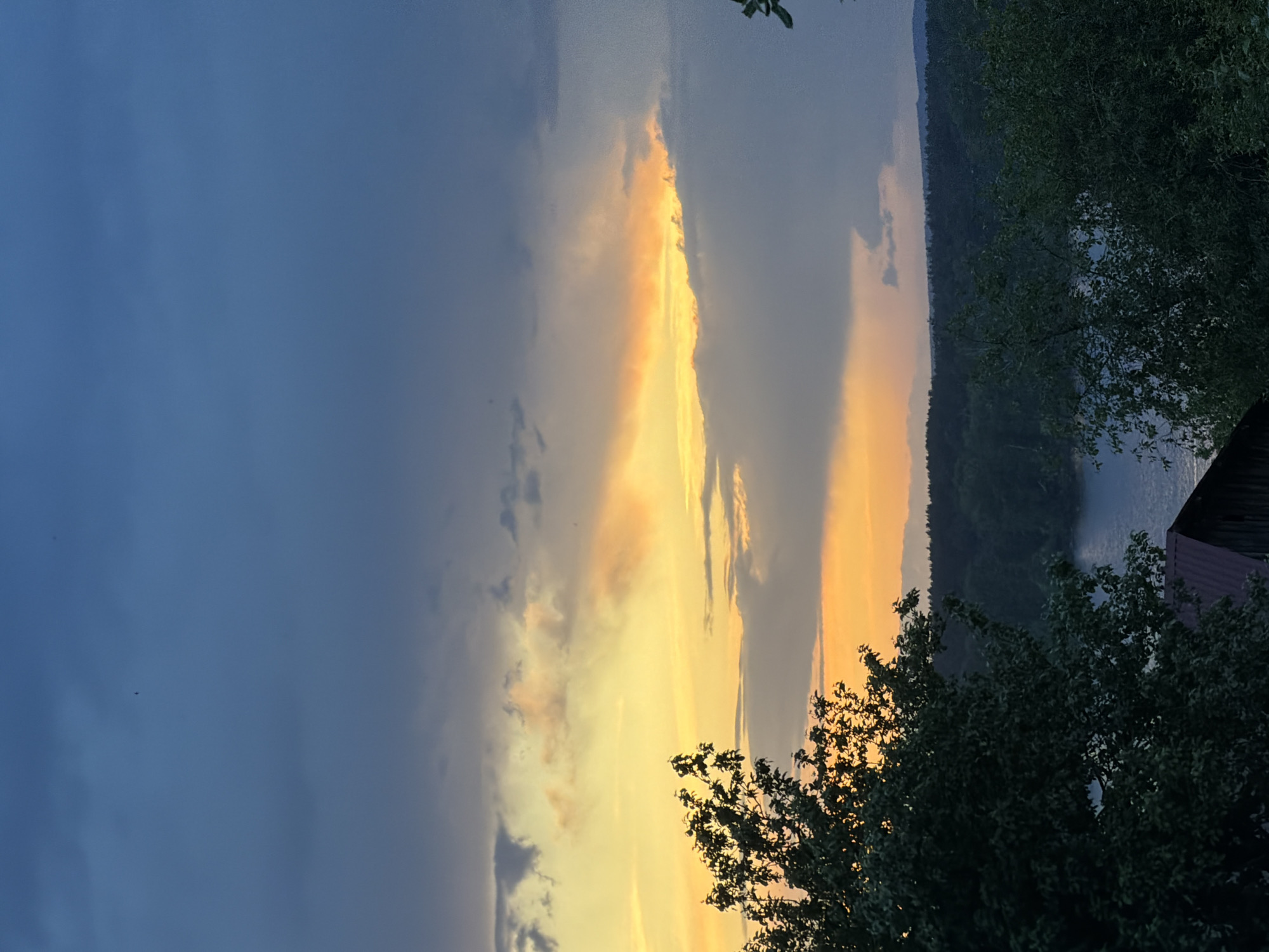 The image is a landscape featuring a body of water, possibly a river or lake, with a shoreline of trees and vegetation. The sky is dramatic with dark clouds transitioning to a bright, golden sunset. Trees are visible in the foreground, obscuring a partial view of a structure in the lower right corner, possibly a building or shed. The location is likely near Chaikovsky, Russia, given the request parameters. The photo captures a moment of natural beauty during sunset, with a contrast between the dark tones of the vegetation and the radiant colors of the sky.
