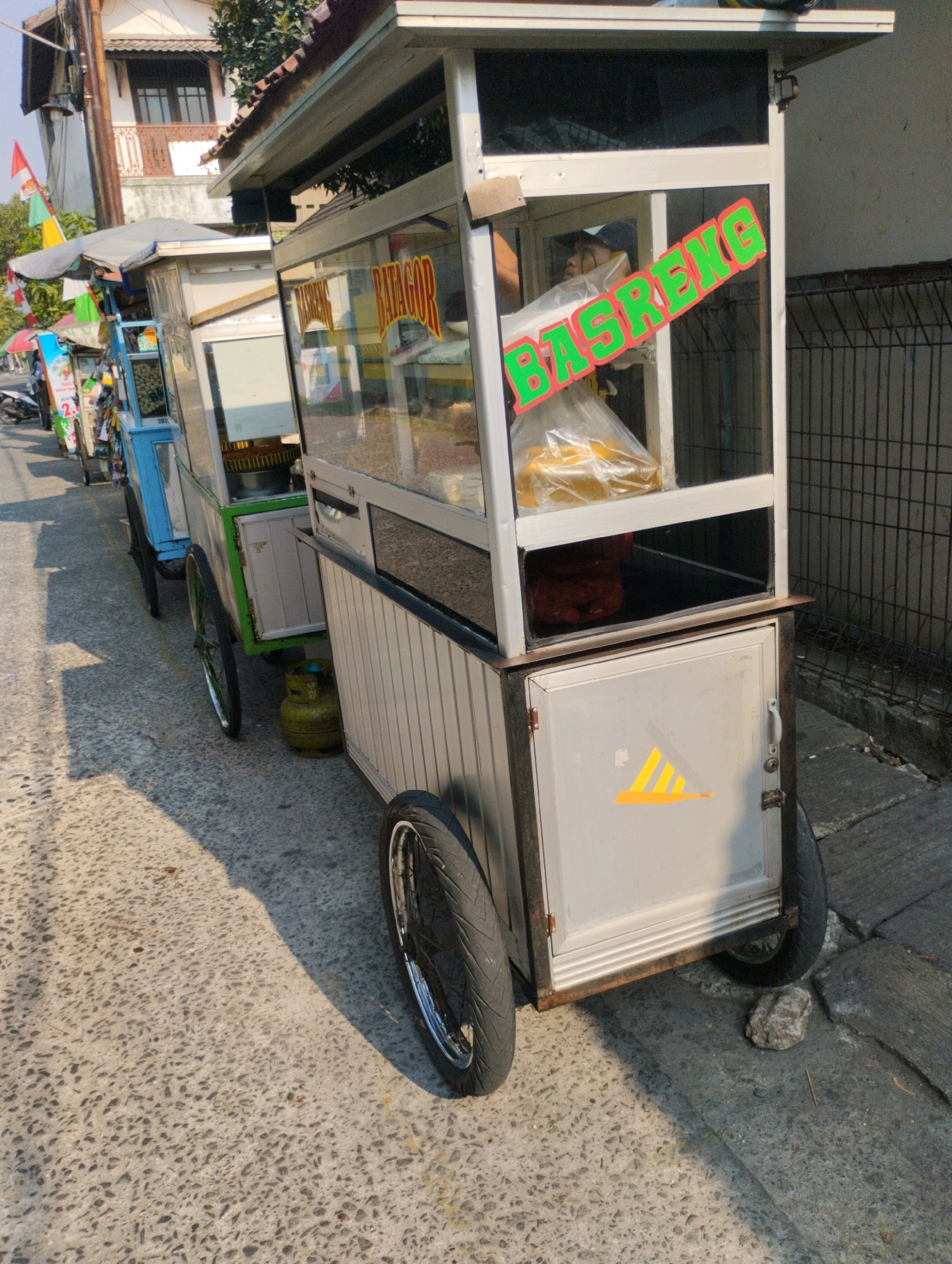 The image shows a food cart selling "BASRENG" (fried fish crackers) parked on the side of a street in Bekasi, Indonesia. The cart is silver with a glass display case and a green sign that reads "BASRENG." There are plastic bags of food inside the display case, and a person, likely the vendor, is visible through the glass. The cart has two large wheels and a smaller wheel in the front. A yellow gas cylinder is placed on the ground next to the cart. In the background, there is a building with a red tile roof and a fence. The street is paved with concrete and has some shadows cast by the sun. Several other food carts are visible further down the street, suggesting a market or a busy street food area. The overall impression is of a typical street scene in Indonesia where mobile food vendors are common.