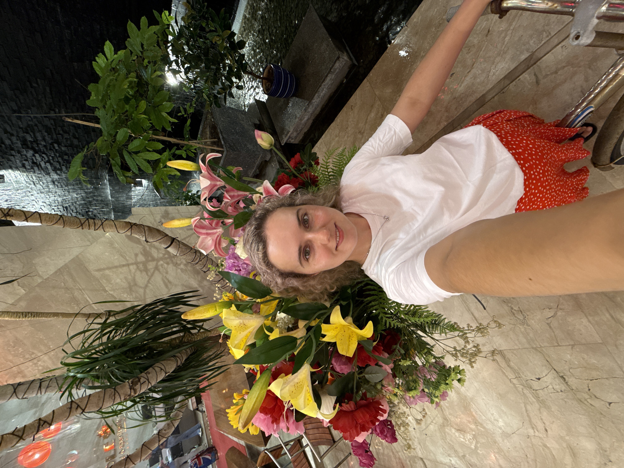 A woman with blonde curly hair wearing a white shirt and red skirt poses with a large floral arrangement. The scene appears to be in Nha Trang Ward, Vietnam, given the unique arrangement and architectural elements visible. A bicycle is also present. A water feature and greenery are in the background.