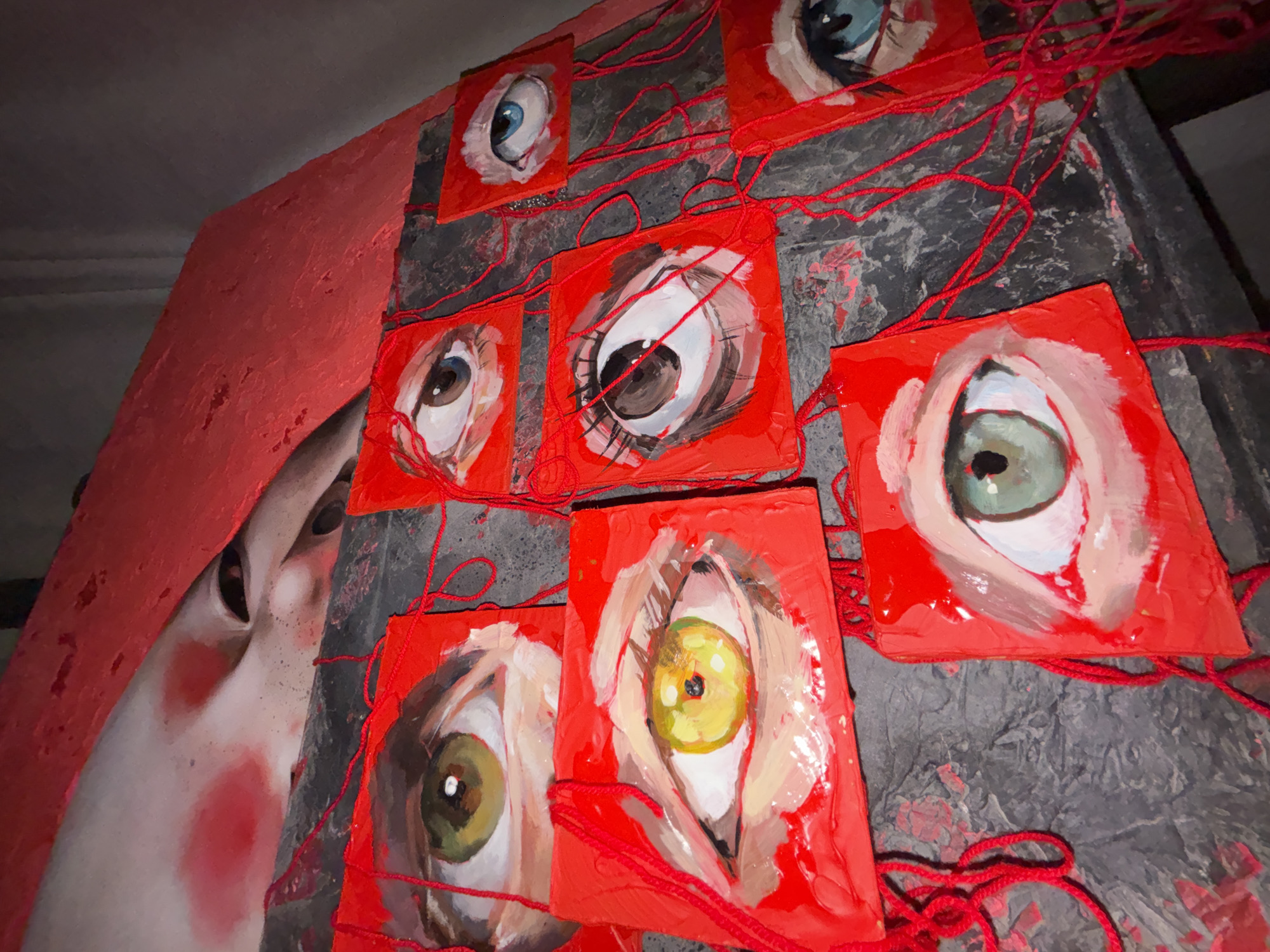 This media file presents an art installation, likely indoors, from Krasnoyarsk, Russia, captured from a low, upward-angled perspective. The central elements are a series of painted eyes and a partial figure, interconnected by red yarn.
Nine small, square-shaped canvases or wooden boards, vibrantly painted in red, are arranged across a dark, textured surface. Each board features a single, expressively painted human eye. The eyes display diverse iris colors, including shades of blue, brown, green, and yellow-gold. Many exhibit visible blood vessels or redness in the sclera, giving them an intense, somewhat unsettling, and highly watchful appearance. The painting style is characterized by visible brushstrokes and a stylized, almost exaggerated realism.
Loops and strands of bright red yarn crisscross and connect these eye paintings, forming an intricate, web-like entanglement across the dark background. To the left of the composition, a large, uniformly red painted wall or panel partially obscures a human-like figure. Only a portion of its face is visible, showing pale skin, prominent rosy cheeks, and dark, upward-gazing eyes. This figure appears to be emerging or peering from behind the red barrier.
The dominant red color, from the canvases and yarn to the large panel, creates a cohesive and impactful visual. The overall impression conveyed by the multitude of watchful eyes and their entanglement, combined with the partially hidden figure, suggests themes of surveillance, interconnectedness, or an omnipresent gaze. There is no visible text in the image.