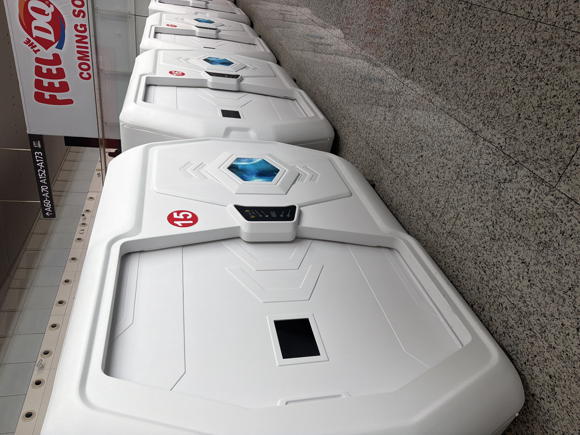 The image displays a row of white, futuristic-looking capsule sleeping pods or rest units in an indoor public space. The perspective is angled, looking along the length of the pods which are lined up against a wall. The floor consists of light grey, speckled granite tiles.
Each capsule is sleek and modern, primarily white with geometric accents. The top of several capsules features a hexagonal, blue-lit display showing a cosmic or planetary image. Below this, a small control panel with various icons (including what looks like Wi-Fi, power, and audio symbols) is visible. The closest pod has a prominent red circle with the number "15" on it, and the next in line shows "16." A small black rectangular window is also visible on the front section of the nearest capsule.
In the background, a large white banner advertising "THE FEEL DQ" (likely Dairy Queen) with the text "COMING SO" (part of "COMING SOON") is prominently displayed. Below this banner, a black directional sign reads "↑A60-A70 A152-A173," indicating gate numbers. Another smaller piece of text, "355-004," is faintly visible on the banner. No people are present, and the artificial lighting suggests an indoor setting, likely an airport or a large transit terminal given the gate information and commercial advertisement.