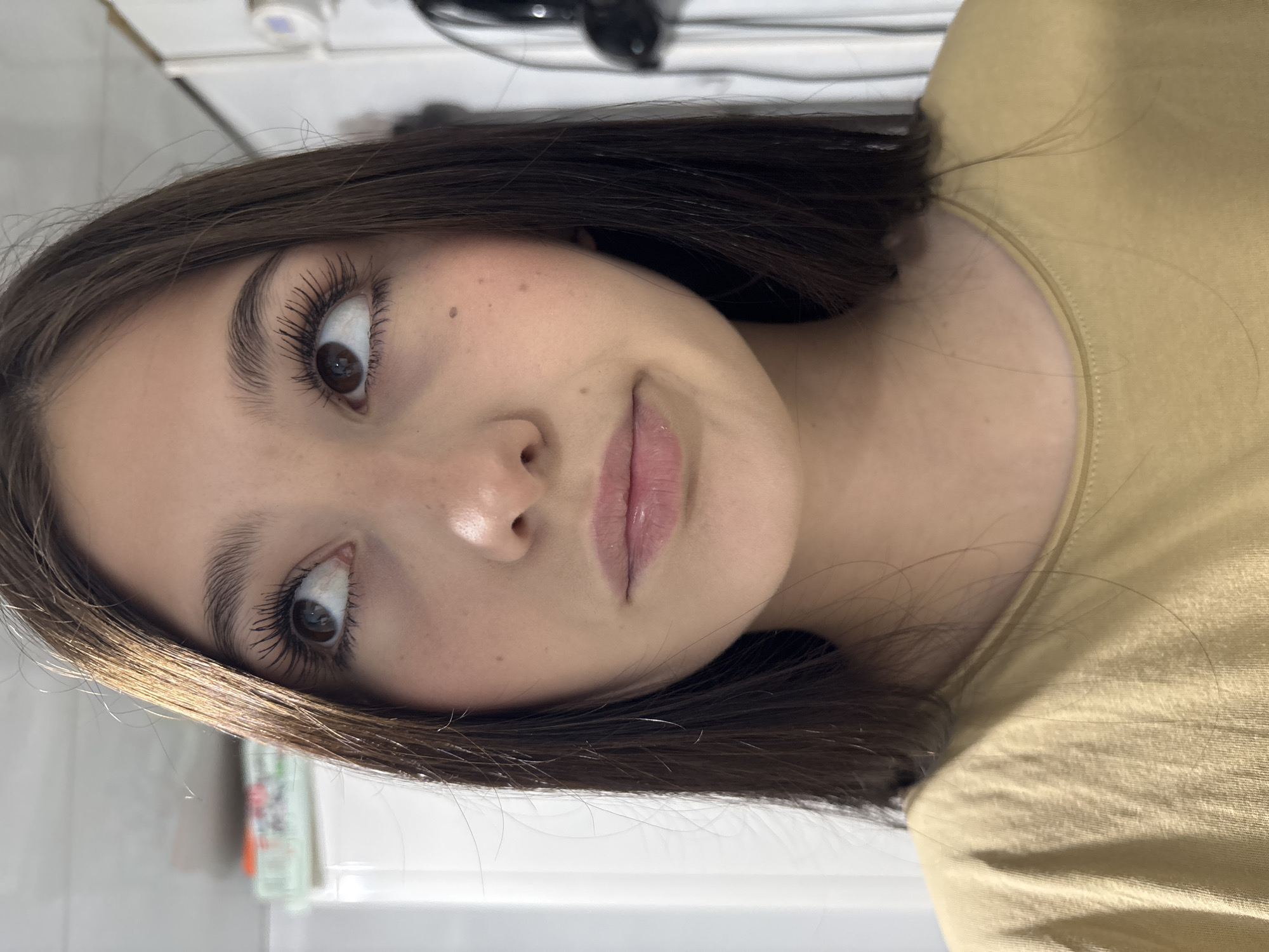A portrait-oriented photo, rotated 90 degrees clockwise, captures a young woman looking directly at the camera. She has dark brown hair, styled casually, and large, dark brown eyes with noticeably long and defined eyelashes, suggesting the use of mascara or extensions. Her eyebrows are well-groomed. Her skin appears clear, with a few visible freckles across her nose and cheeks. Her lips are a light pink or nude shade, slightly parted. She is wearing a simple, light-colored (possibly tan or light yellow) t-shirt with a round neckline.
Her expression is wide-eyed, perhaps with a hint of surprise or curiosity, and her lips are slightly pursed, giving a soft, natural look. The background is softly blurred, showing an indoor setting with various light-colored surfaces and some indistinct objects, including what might be a dark electronic device (possibly a hairdryer) and white shelves or tiles. The lighting suggests a bright, indoor environment, likely daytime.
No decipherable text is visible in the image. The photo provides a close-up view of her face, highlighting her features and makeup. The location context is Фоки, Russia.