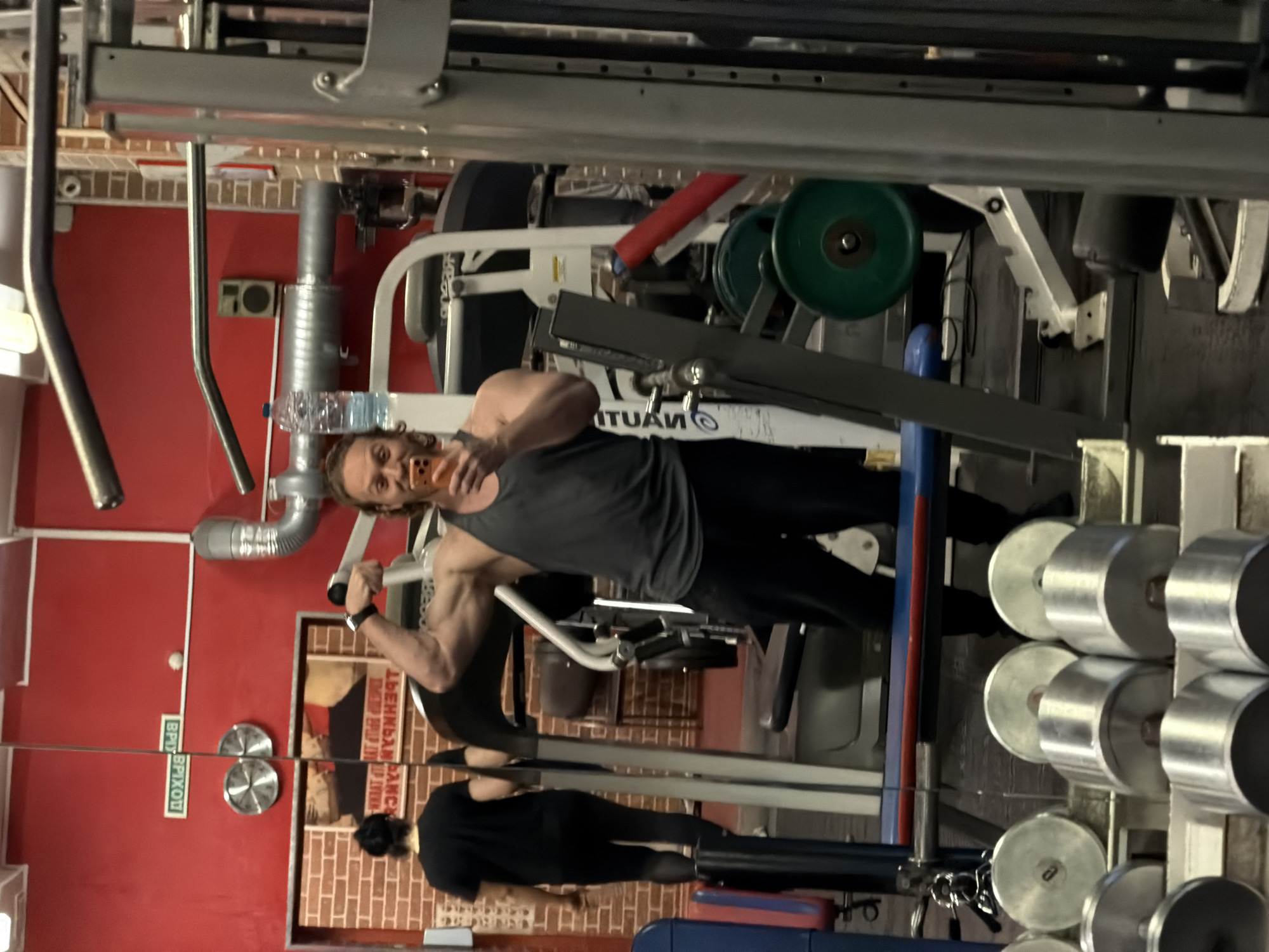 The image depicts a scene within a gym, possibly in Saint Petersburg, Russia, based on the signage visible. The primary subject is a man in workout attire, operating a weight machine. He is holding a smartphone and appears to be taking a photo or video of himself. Another person is seen walking on a treadmill in the background. The gym contains various exercise equipment including weight machines, dumbbells, and weight plates. The interior walls are painted in red.