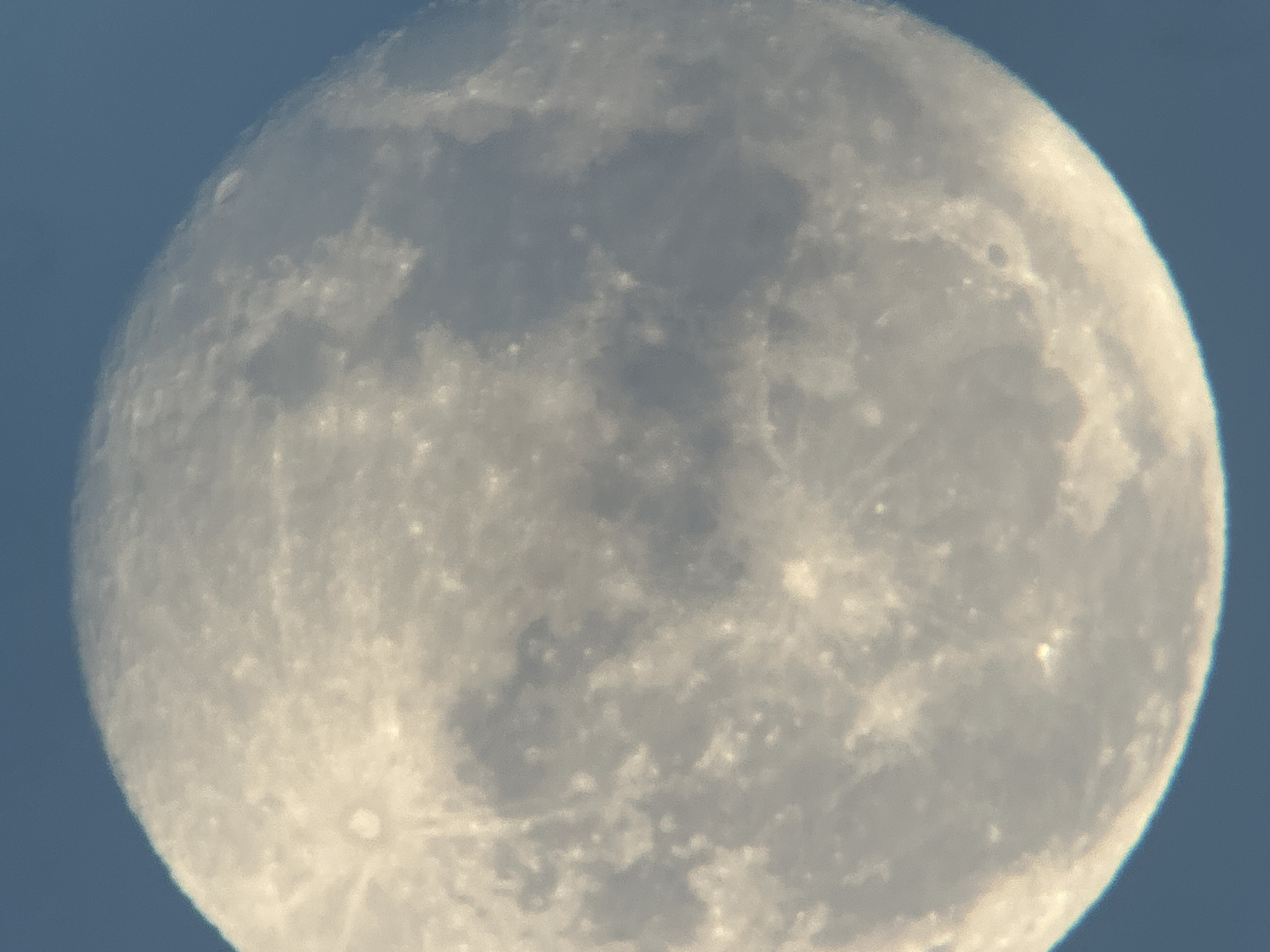 A close-up, slightly blurry photograph shows the moon against a clear blue sky. The moon is large and fills most of the frame, displaying visible craters and darker maria (lunar plains). The lighting suggests it is daytime or twilight. There are no identifiable landmarks or context to place this image in Lyubertsy, Russia, or any other specific city.
