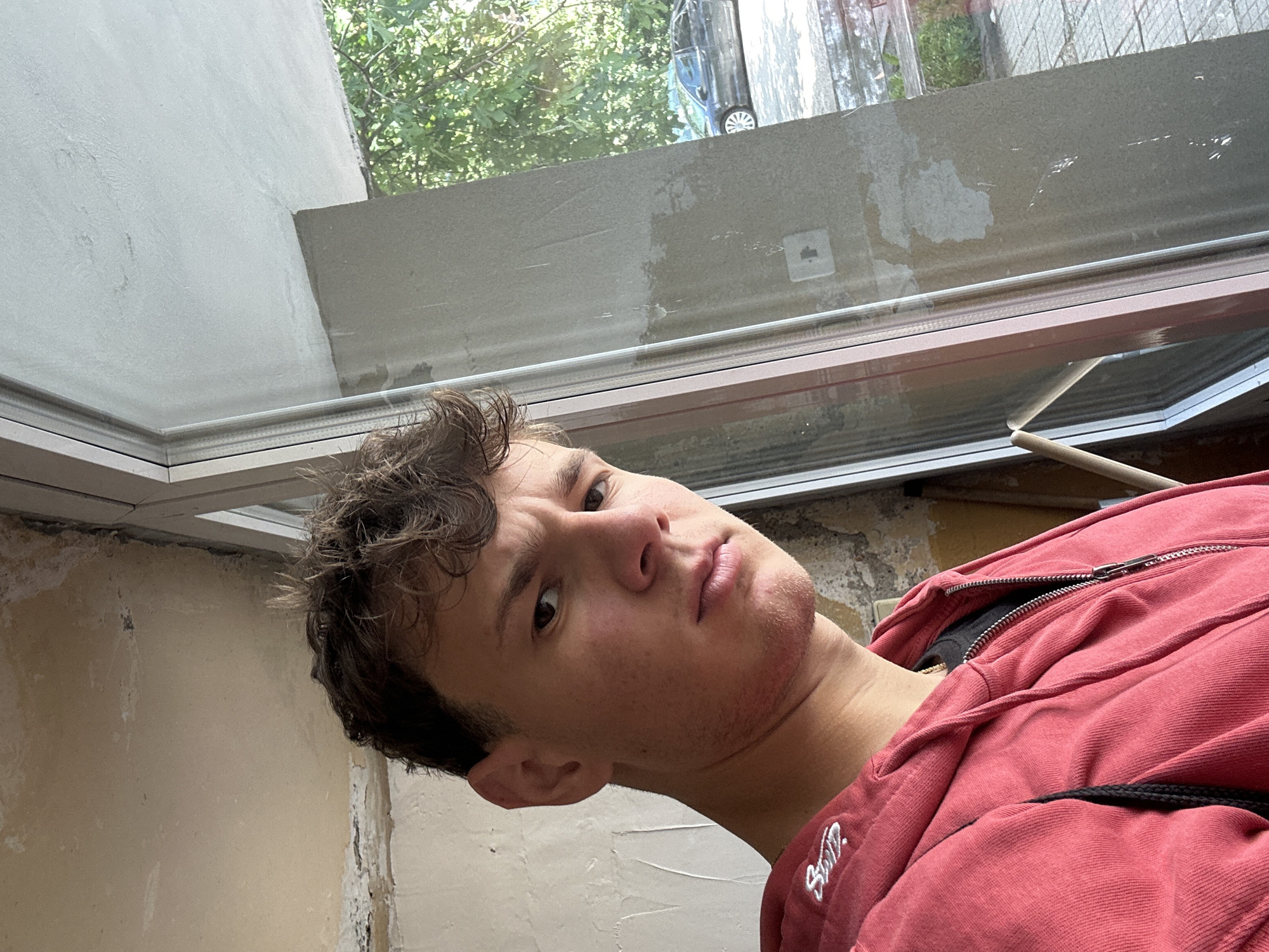A head-and-shoulders selfie depicts a young man with brown, curly hair and a red zip-up hooded sweatshirt. He is positioned indoors, near a window with exterior views of greenery and a vehicle. The wall adjacent to the window exhibits signs of construction or renovation, with exposed patches and uneven surfaces. The location is indeterminate and not confirmed to be Elhovo, Bulgaria, due to a lack of contextual details.