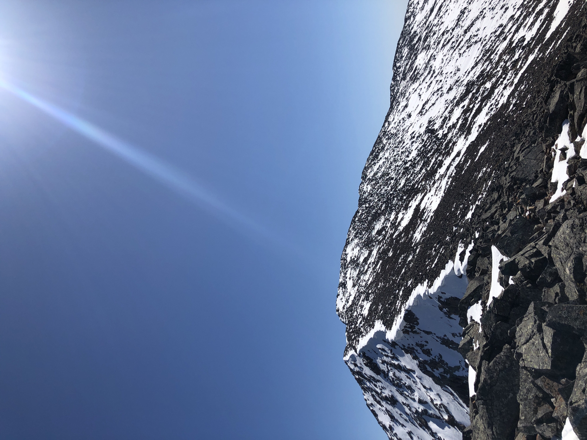The image depicts a steep mountainside, partially covered in snow, juxtaposed against a clear blue sky with a lens flare from the sun. The mountainside consists of dark rocks interspersed with patches of white snow. The sky is a gradient of light blue, with the sun's glare visible in the upper left corner. The image is rotated, presenting a somewhat unusual perspective. There is no indication of location. The city and country are unknown.