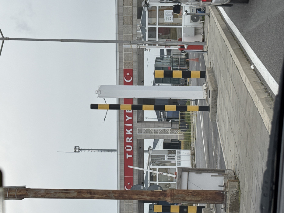 The image depicts the Turkish entry point of the Lesovo-Hamzabeyli border crossing, geographically associated with Lesovo, Bulgaria. The vantage point is from inside a vehicle positioned in a queue lane.
The scene is dominated by a grey overhead archway spanning multiple lanes. A prominent red band on the archway displays "TÜRKİYE" in white capital letters, flanked by two Turkish flag emblems (white star and crescent on red). The archway's supports feature a repeating grey geometric pattern.
Multiple black and yellow striped bollards delineate vehicle lanes. A tall, white, rectangular structure is centrally positioned in one lane, flanked by these bollards. To the left, a white-framed glass guard booth is visible with its white barrier arm in a raised position, and yellow metal railings are behind it. To the right, another white-framed glass guard booth, partially visible, has an external air conditioning unit. An orange traffic cone stands near this right booth.
In the immediate left foreground, a rusted brown metal pole rises vertically. A tall, grey metal street light pole is on the right side of the frame. In the background, a tall, grey lattice-style communication tower is visible above the archway.
The roadway is asphalt with white lane markings. A grey concrete slab sidewalk borders the right side of the road. A small white piece of litter is visible on the ground next to the sidewalk curb. The rear of a white sedan is partially visible on the far right. The sky is uniformly overcast and light grey. No people are discernible in the frame.