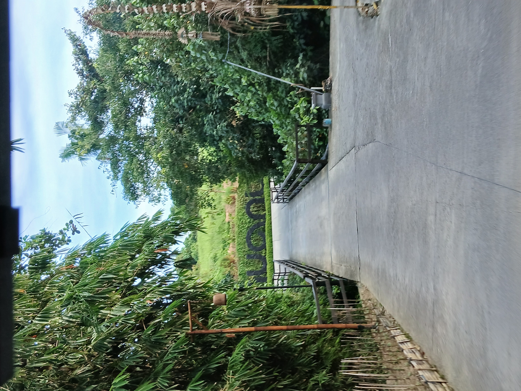 The image captures a paved pathway leading towards a bridge. The pathway is flanked by lush green foliage on the left and a variety of trees and vegetation on the right. In the background, a grassy hill rises, and a large, dark, three-dimensional sign spelling out "UYOON" is visible. The sign is partially obscured by the surrounding greenery. The scene appears to be an outdoor recreational area or resort, possibly part of the UYOON resort in Kediri, Indonesia, given the presence of the resort's sign and the tropical vegetation. The sky is a bright blue with scattered clouds.