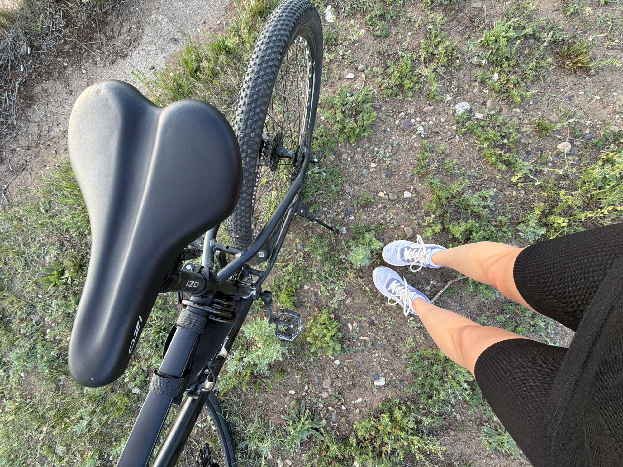 An overhead shot captures a person standing on a dirt path next to a black bicycle. The person is wearing black cycling shorts and light blue athletic shoes. The bicycle is positioned at an angle, with its black seat prominently displayed. The ground is covered in dirt, small rocks, and sparse green vegetation. The background consists of more of the same natural terrain. There is no visible indication of the location within Chaikovsky, Russia.