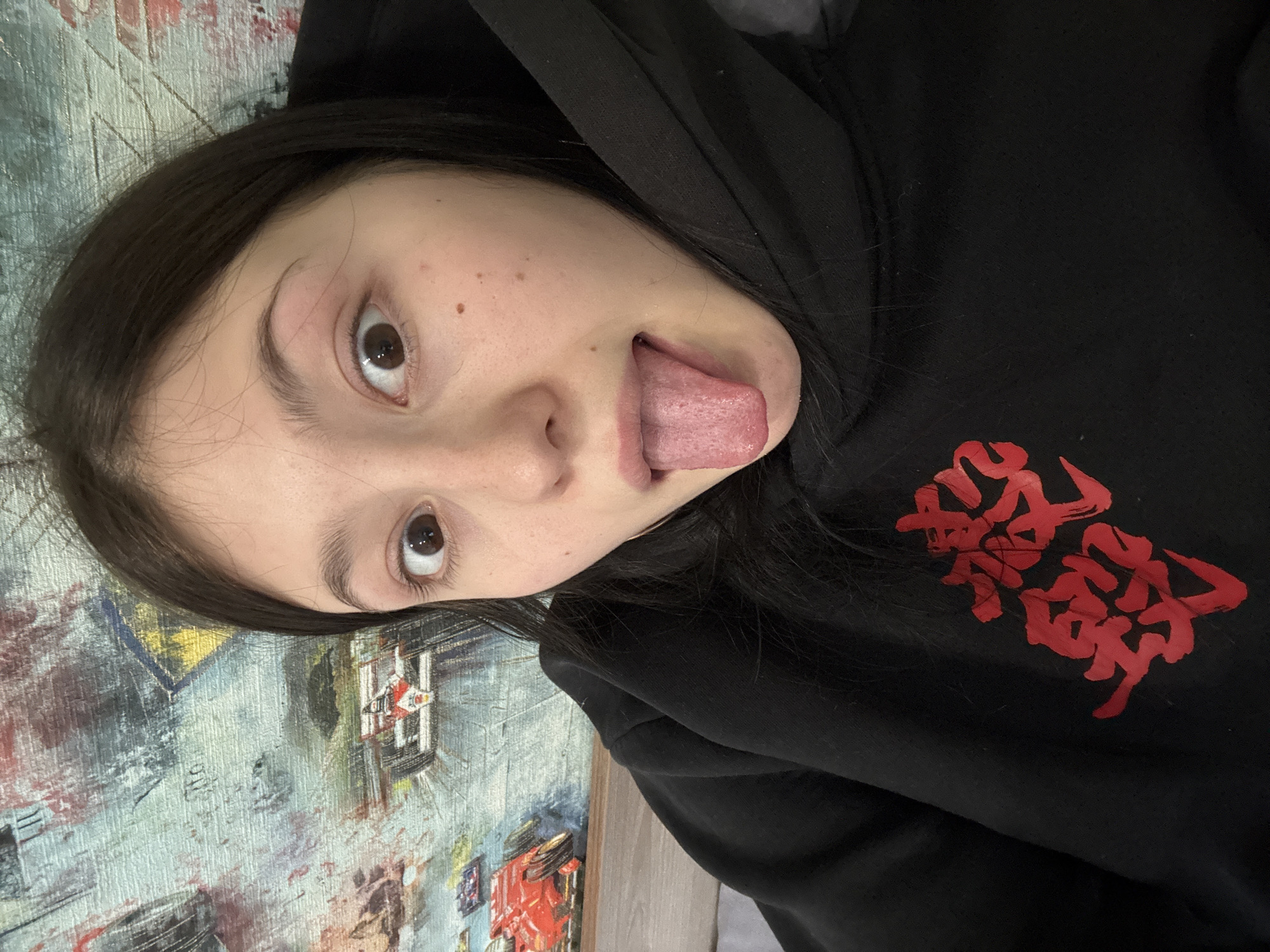 A young person lies on their back, sticking out their tongue. They wear a black hooded sweatshirt with red Japanese-style script on the front. The background appears to be a wall or surface with abstract art. The image's orientation is tilted. The location Фоки, Russia is not ascertainable.