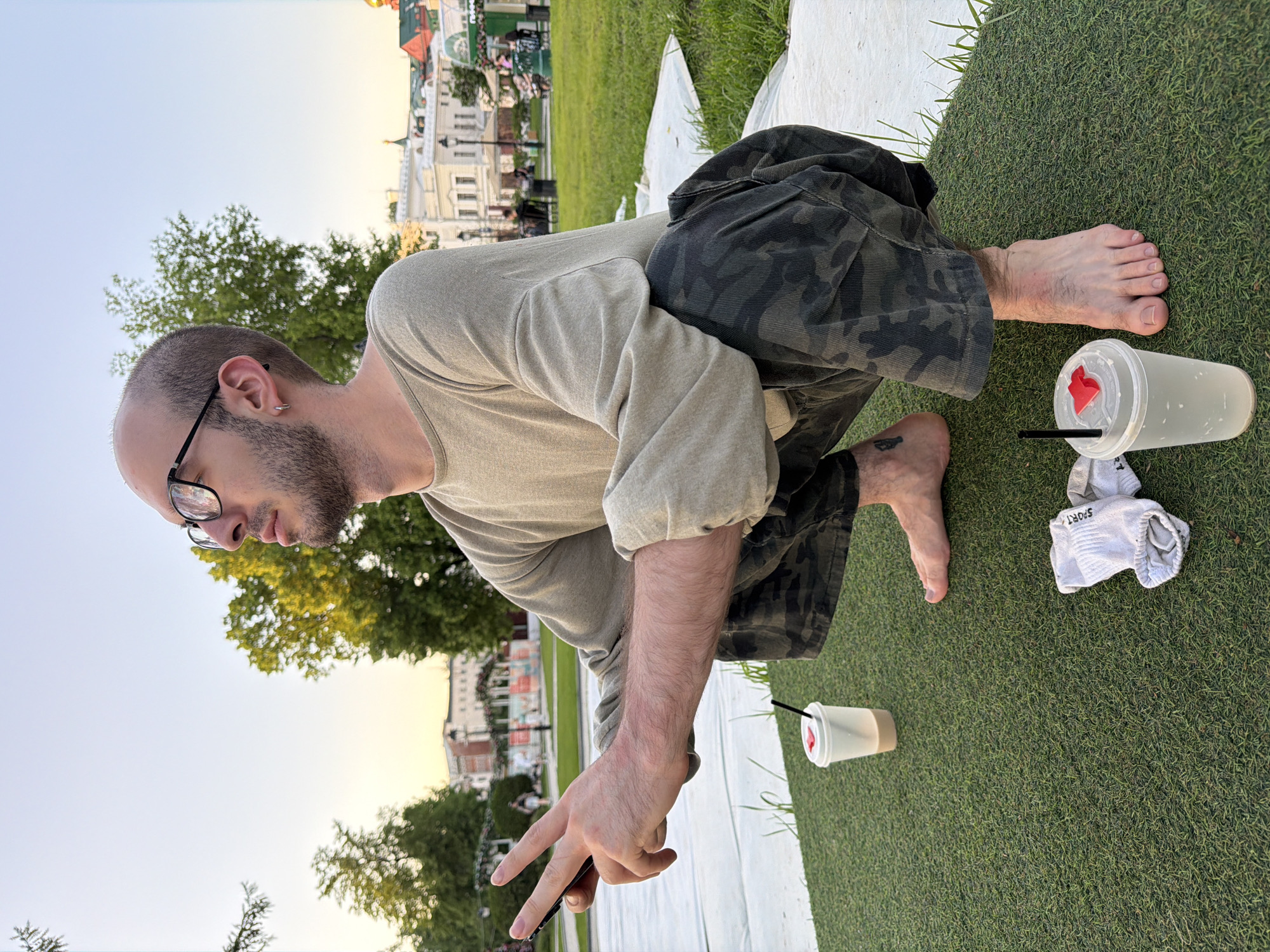 The image depicts a man squatting on green grass next to a white surface. He is wearing glasses, a light brown long-sleeved shirt, and camouflage shorts. He is holding a cell phone in his left hand, making a "peace sign" with his fingers. To his right, there are two clear plastic cups with lids and straws, along with a pair of white socks. In the background, there is a white building and greenery, suggesting a park or outdoor setting. This is likely the city of Moscow, Russia.