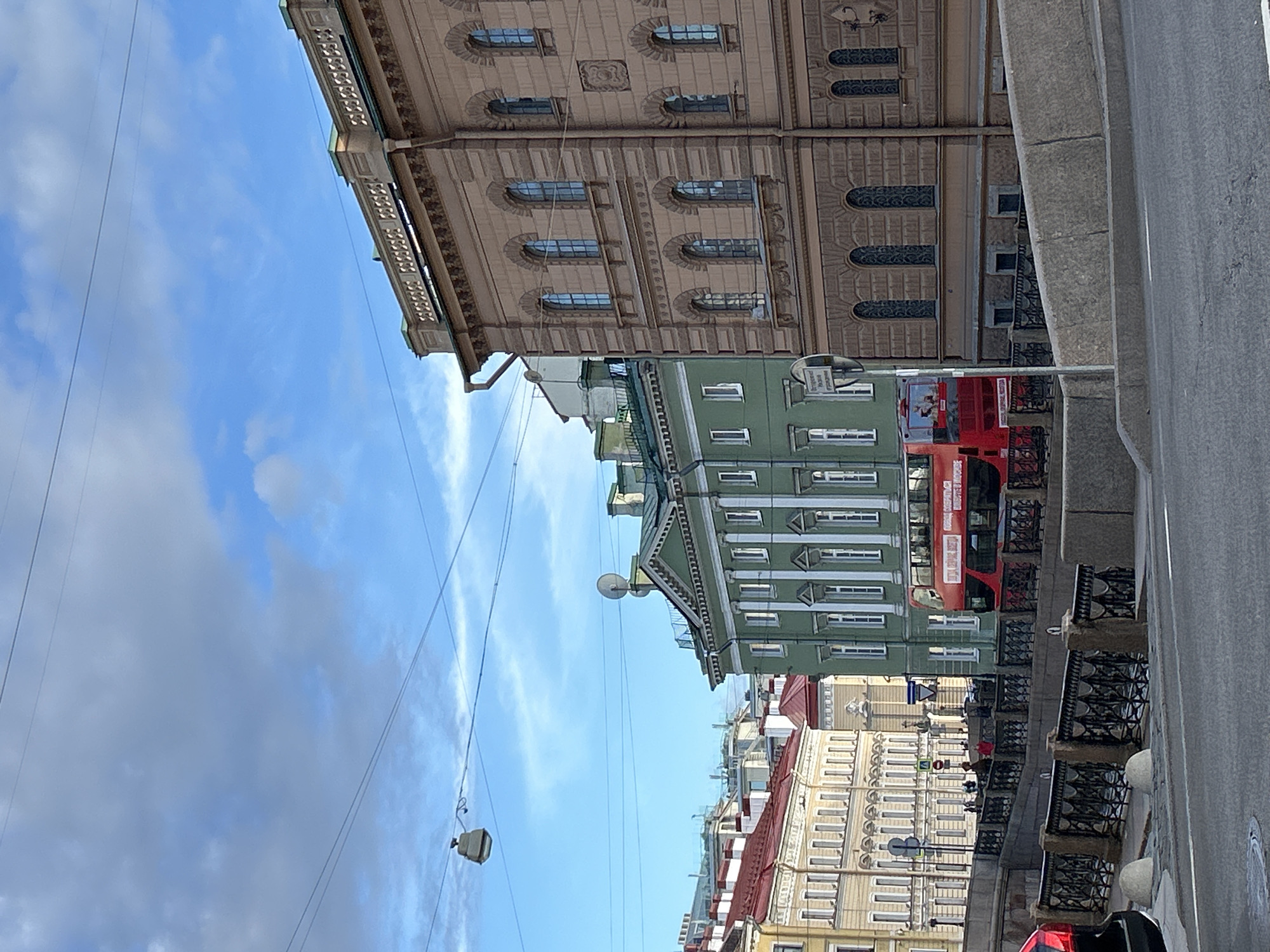 This media file depicts a street scene in an urban setting, likely a European city given the architecture of the buildings. The image is oriented vertically, with the sky at the bottom and the buildings extending upwards.
The primary subjects are several buildings with ornate facades and numerous windows. The buildings vary in color, with one prominent green building and a lighter beige building with detailed ornamentation. A bright red double-decker bus is visible parked in front of the green building, suggesting a tour bus or public transport. A few people can be seen on a sidewalk or promenade in front of the buildings, some appearing to be tourists.
Notable details include the architectural styles of the buildings, which suggest a historical or classic design. The sky is blue with scattered white clouds, indicating it is daytime. The presence of power lines and a lamppost adds to the urban feel. A traffic sign is visible near the red bus. There is a stone balustrade along what appears to be a waterway or canal next to the street.
The scene appears calm and the weather pleasant for walking and sightseeing. No specific emotional tone is strongly conveyed, but the image suggests a typical day in a well-trafficked urban area. There is some text visible on the traffic sign and on the side of the red bus, but it is not legible enough to be transcribed. The overall impression is of a picturesque city street.