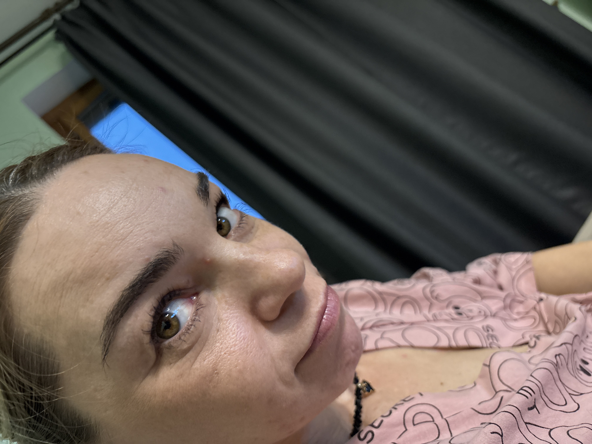 The image shows a close-up of a woman's face, cropped from the nose up, with her head tilted to her left. She has brown eyes, dark eyebrows, and light-colored skin with some texture visible. She is wearing a pink shirt with a black choker necklace. The background is mostly out of focus, with a dark curtain on the right and a bright blue area on the left. There is no information in the image to suggest a location in Sinyavino, Russia.