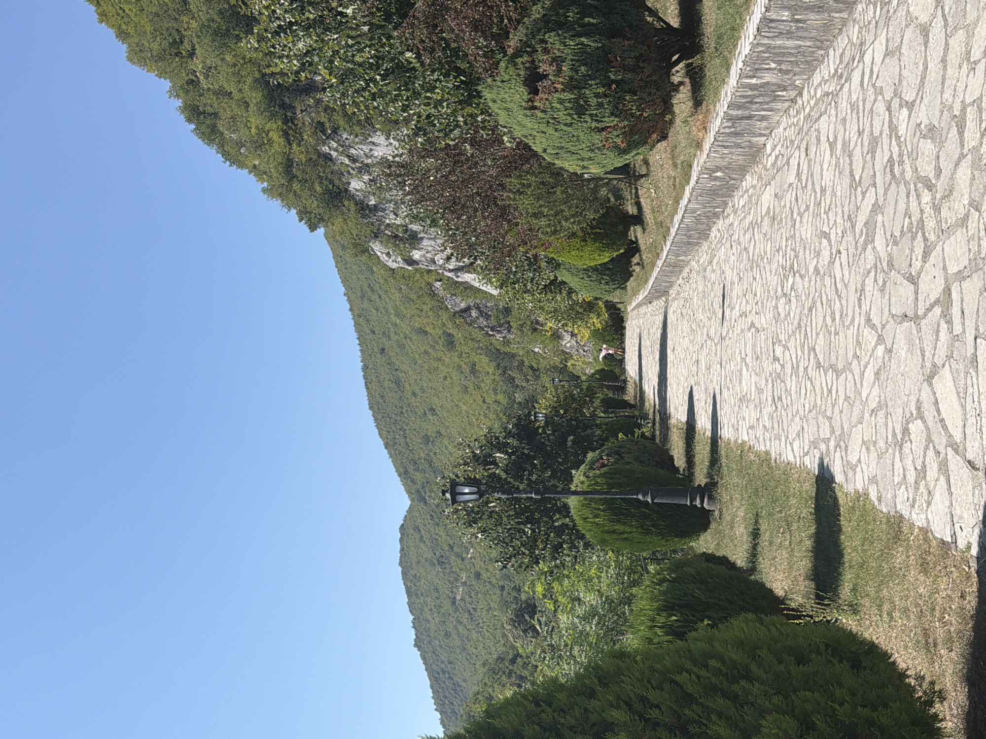 A stone pathway lined with manicured trees and shrubs leads uphill towards a forested mountain range under a clear blue sky. A vintage lamppost stands on the left side of the path. In the distance, a person in pink shorts and a white top walks away from the camera. The scene appears to be at Manastir Morača, Montenegro.