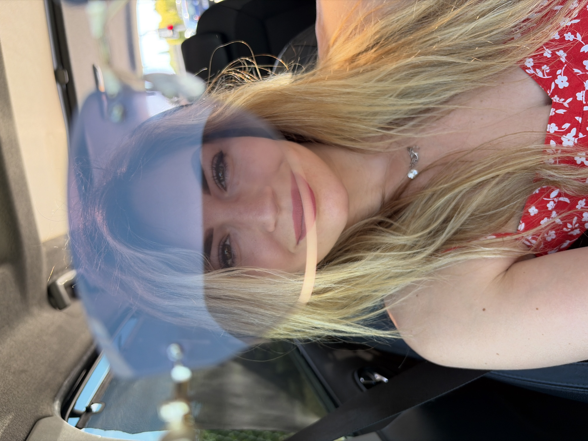 The image presents a close-up, sideways portrait of a young woman with long, wavy blonde hair, captured within a vehicle. She is smiling softly, revealing light-colored eyes defined by makeup and pink lipstick. Her fair skin shows a scattering of freckles or moles, particularly on her arms and neck. She wears a red top or dress with a white floral pattern and a silver necklace adorned with a clear stone pendant.
The setting is the interior of a car, with dark upholstery and a seatbelt visible. The photo is taken through glass, creating prominent reflections that overlay the woman's image. A significant light blue or purple reflection covers parts of her face and hair, hinting at the color of the car's bodywork or another object. Blurred reflections of an outdoor environment suggest a bright, sunny daytime setting, with hints of green and yellow foliage.
The woman appears relaxed and content, indicated by her gentle smile. There is no visible text in the image. The unique angle and layered reflections give the photo a dynamic and intimate feel, placing the woman inside a moving or stationary vehicle in Podgorica, Montenegro.