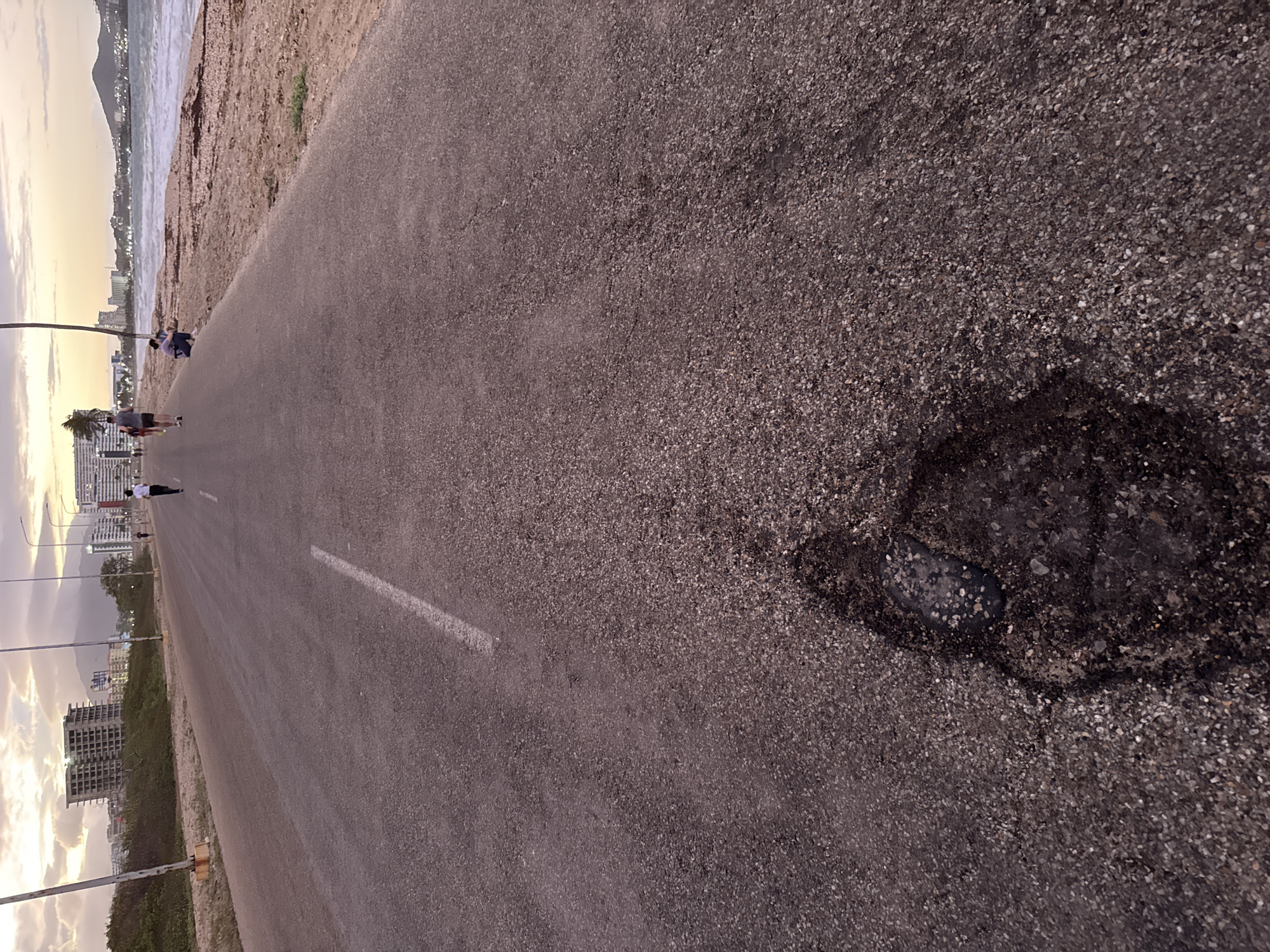 An asphalt road stretches into the distance, with a white line visible on its left edge. On the right side of the foreground, a prominent, dark pothole or patch is visible on the road surface. The road appears to run parallel to a coastline, with a sandy beach or shoreline leading to a body of water (likely the sea) on the right. In the distance across the water, mountains are visible under a pastel sky.
Several people are present along the road and near the water. A person in a white shirt and dark pants walks away from the viewer further down the road. Closer to the water, a group of individuals are standing and sitting on the beach or next to the road. On the left side of the road, numerous lampposts line the path, and in the background, various buildings and urban structures can be seen.
The lighting suggests either dawn or dusk, with soft, warm hues in the sky indicating late afternoon or early morning. The overall atmosphere appears calm and peaceful. The scene depicts a coastal promenade or road in an urbanized area, with the given context placing it in Porlamar, Venezuela. No visible text is present in the image.