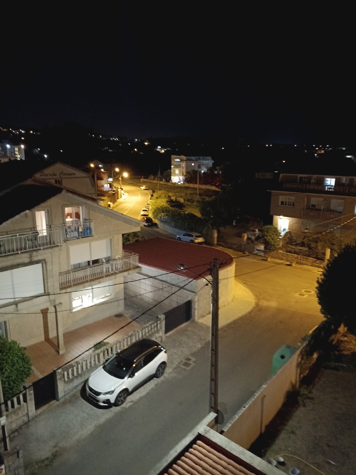 The image depicts a nighttime aerial view of a residential area in A Lanzada, Spain. In the foreground, a white car with a black roof is parked on a paved driveway next to a two-story house with illuminated windows and balconies. The house has a light-colored facade, white-framed windows, and a dark roof. A black metal fence borders the driveway.
Further into the scene, a winding street is visible, illuminated by streetlights, with several cars parked along its edge. More houses and buildings, some with lit windows, are scattered throughout the landscape, extending into the background. Trees and vegetation are present, adding to the suburban setting. The sky is dark, indicating nighttime. The overall impression is a quiet residential neighborhood at night.