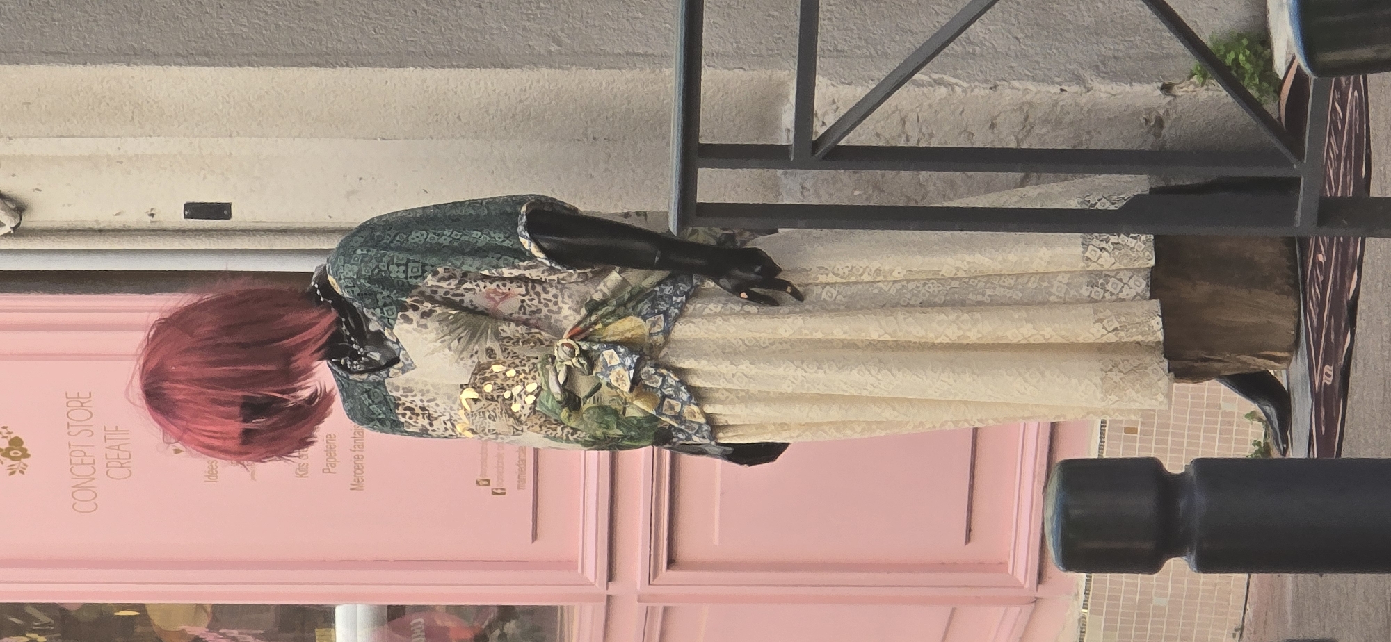 A full-body mannequin is displayed in front of a pink storefront. The mannequin has bright red, messy hair and is wearing a dark beaded necklace. Its outfit consists of a patterned V-neck top with green, beige, and animal print accents, and a long, cream-colored lace skirt. The mannequin's arms and hands are black, with one hand resting on its hip and the other extended forward, fingers slightly curled. The storefront has text that reads "CONCEPT STORE CREATIF" in a light orange font. Below this, smaller text lists items like "Idées," "Kits," "Papeterie," and "Mercerie fantaisie." A metal railing runs in front of the mannequin and the store. The ground in front of the mannequin has a dark rug with a light pattern. The mannequin stands on a large brown, wooden base. The background shows a textured, light-colored wall.