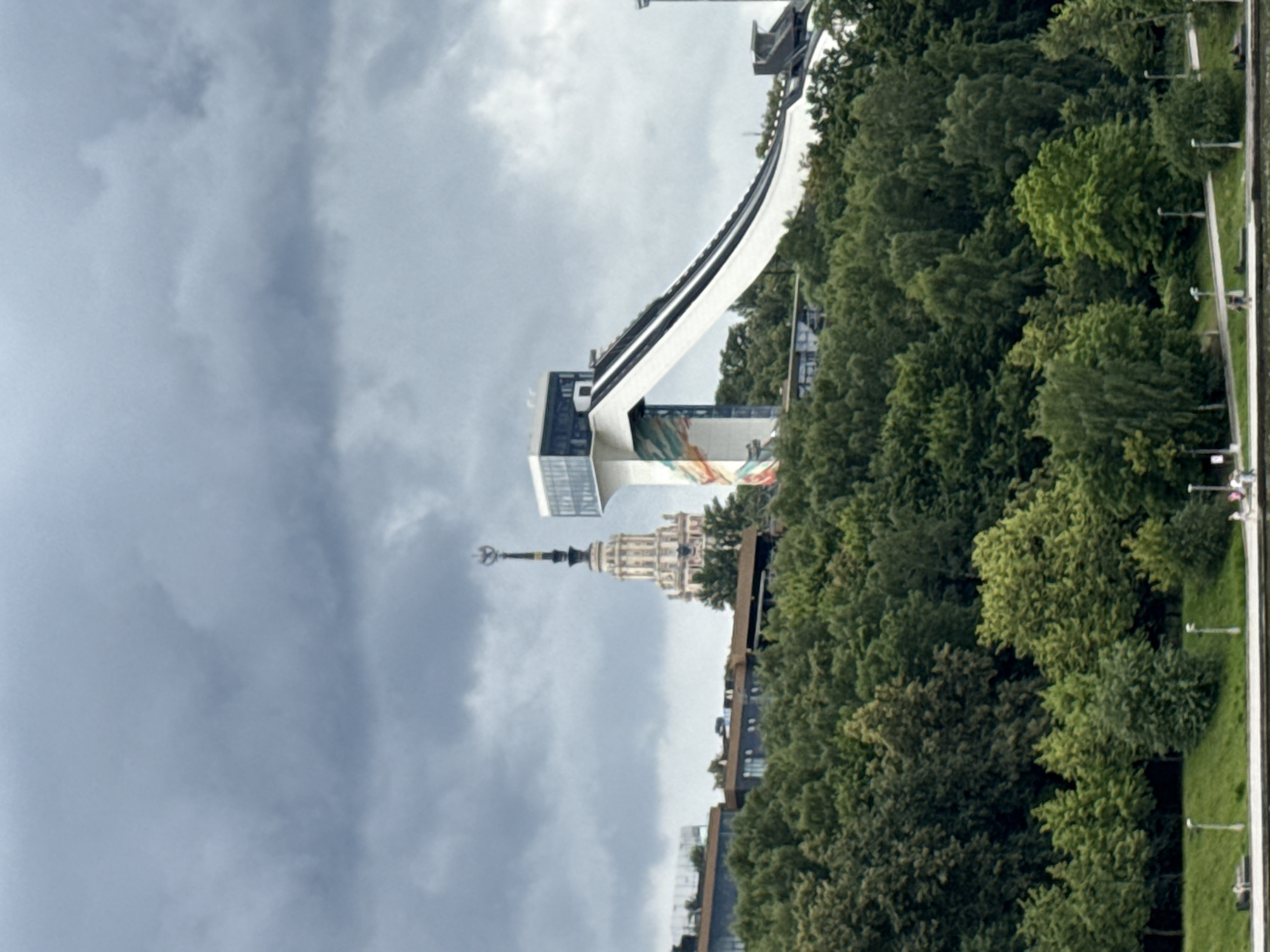 The image captures a scene in Moscow, Russia, featuring a ski jump facility atop a hill, with a distinctive Stalinist skyscraper visible in the background. The ski jump is a modern structure with a long, white ramp that curves downwards. The lower portion of the ramp features decorative colorful patterns. Above the ramp, there is a glass-enclosed structure.
Behind the ski jump, a tall, ornate Stalinist skyscraper with a spire stands prominently. Its facade is light-colored with multiple tiers and decorative elements. In front of these structures, a dense forest covers the hillside, with varying shades of green indicating a variety of trees and foliage. Some modern buildings with dark facades and flat roofs are partially visible peeking through the trees.
The sky is overcast with heavy, dark clouds, suggesting a somber or potentially rainy day. In the foreground, a body of water, likely a river or canal, is visible. Along the embankment on the far side of the water, a grassy slope is lined with lampposts. A few small figures of people can be seen walking along the path on the embankment. One person is also visible sitting on a bench near the water's edge. The overall impression is of an urban landscape with a mix of natural elements and modern and historical architecture.