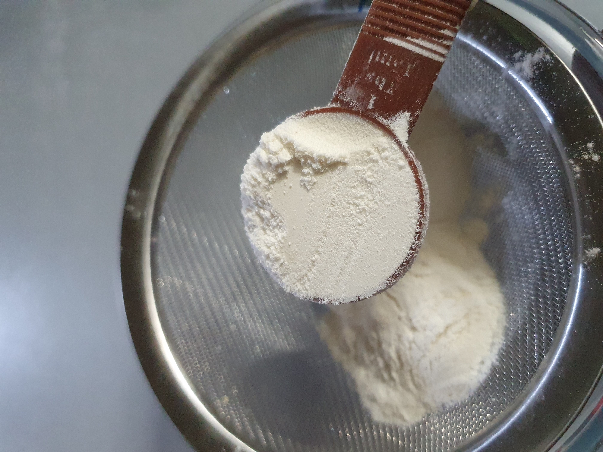 An overhead shot shows a close-up of a measuring spoon filled with white powder, likely flour or a similar ingredient. The spoon is positioned over a fine-mesh sieve, through which more of the white powder is visible in the background. The sieve is resting on a surface that is out of focus but appears to be a kitchen counter or similar setting. The lighting suggests an indoor environment. There are no people or clear indications of the city of Shiraz, Iran.