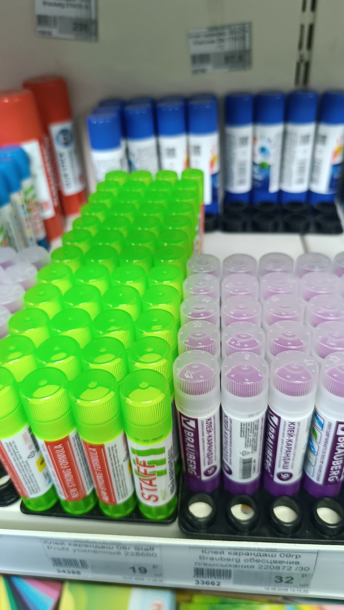 The image displays a shelf in a retail store, likely a stationery or office supply store. The shelves are stocked with rows of glue sticks. In the foreground, on the left, are several bright green glue sticks with white labels that read "STAFF" and "NEW STRONG FORMULA." To the right of the green glue sticks are rows of purple-topped glue sticks with white labels. Some of these labels are partially visible, showing branding such as "BRAUBERG" and "КЛЕЙ-КАРАНДАШ" (glue stick in Russian). Behind the rows of glue sticks, on higher shelves, are more glue sticks in blue and red packaging. The background shows the retail environment with additional shelves and product packaging. Below the products, price tags and product information labels are visible. One label reads "Клей карандаш 08г Staff Profit усиленный 228660" and another reads "Клей карандаш 09гр Brauberg обесцветив." The presence of Russian text on the labels indicates the location is likely in a Russian-speaking country. Given the specific mention of "Chaikovsky, Russia" in the prompt, it's probable this scene is within a store in that city, though the image itself does not contain direct visual evidence to confirm this specific location. No people are visible in the image. The scene is clean and organized, typical of a retail display.
