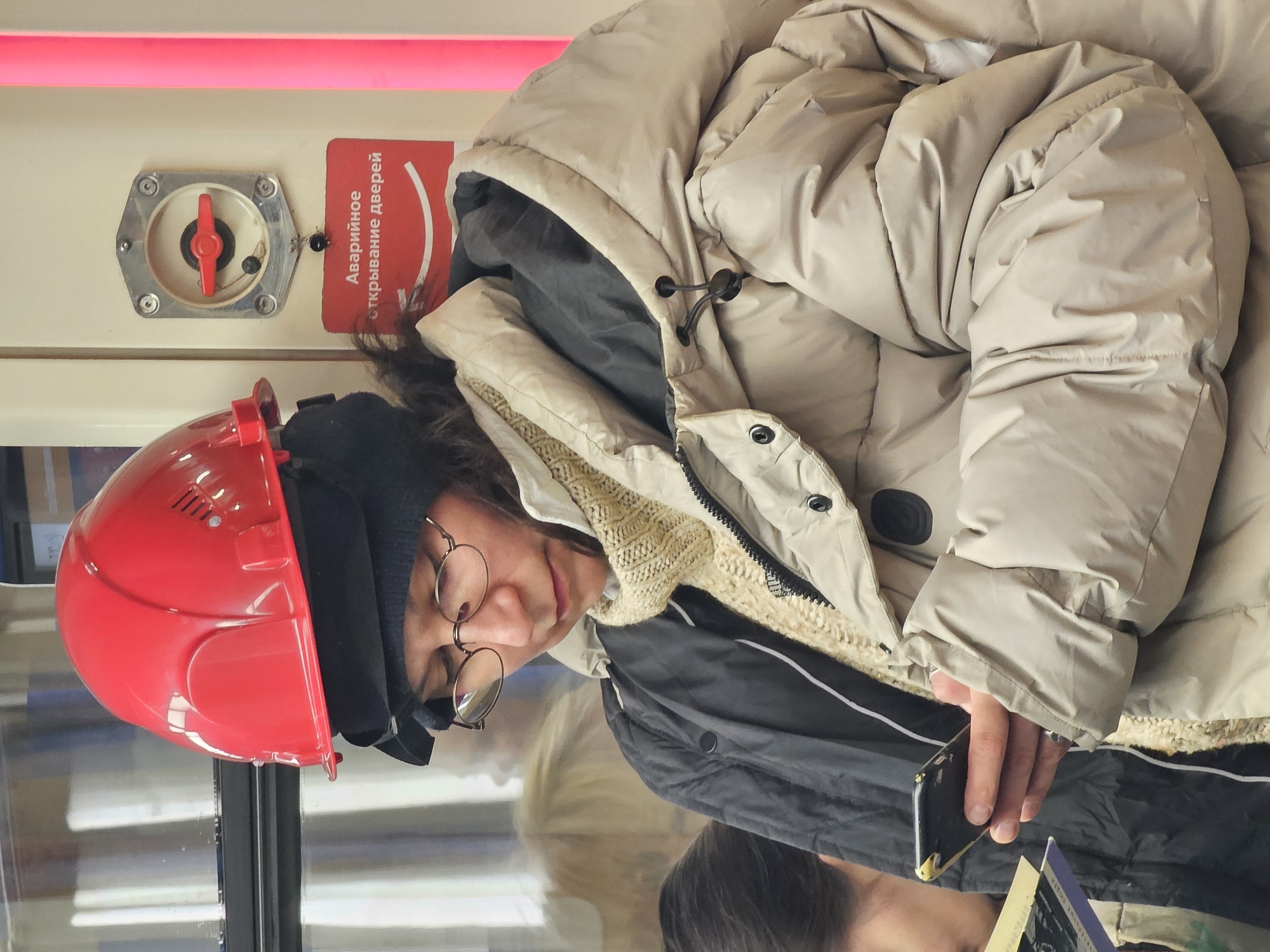 The image captures a person inside what appears to be a metro train or similar public transport vehicle, likely in Moscow, Russia. The main subject is a person, possibly female, with dark, wavy hair, wearing a black beanie and distinctive round, wire-rimmed glasses. She is dressed in a thick, light beige puffer jacket over a black inner layer, suggesting cold weather. A bright red hard hat is perched on top of her beanie.
Her gaze is directed downwards, focused on a black smartphone held in her right hand. A book or magazine is partially visible beneath the smartphone. In the background, mounted on a light-colored wall, is a silver hexagonal emergency mechanism with a prominent red lever. Attached to this mechanism is a red sign with white text. The Russian inscription reads "Аварийное открывание дверей," which translates to "Emergency door opening," confirming the Russian context for the public transport setting. Above this mechanism, a bright red light strip runs horizontally along the top of the carriage wall.
A reflection of another person, possibly with blonde hair, is visible on a reflective surface or window below the main subject, further indicating an indoor public space. The person appears calm and focused on her device, seemingly commuting or traveling.