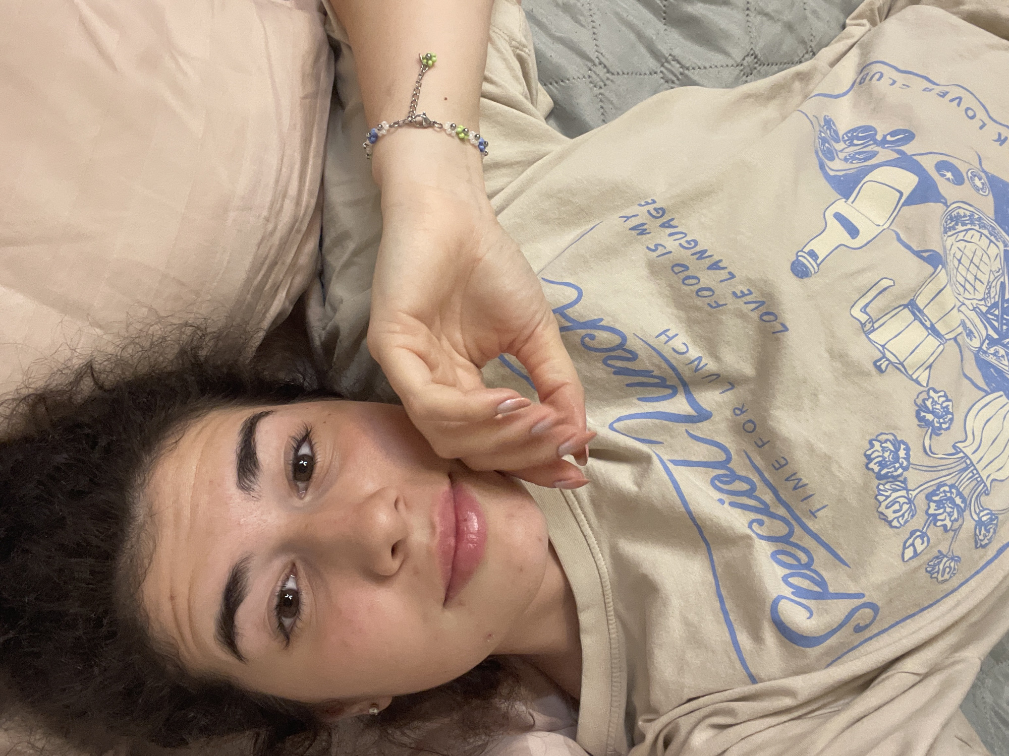 The image is a close-up, overhead shot of a young woman lying in bed. She is looking directly at the camera with her head resting on a pale pink pillow. Her hair is dark and curly, framing her face. She is wearing a light-colored t-shirt with blue text and graphics. The text on the shirt reads "Special Lunch" and "Food is my love language." There is also a graphic of a moka pot and a bottle. A beaded bracelet with blue and green beads is visible on her left wrist. The background consists of pillows and bedding in muted tones. The lighting appears to be artificial, casting soft shadows. There are no visible elements in the image that indicate a specific location in the city of Sofia, Bulgaria.