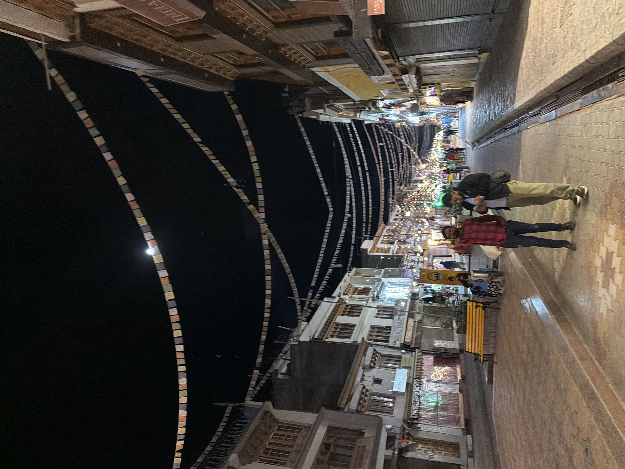 The image captures a nighttime scene on a street in the city of Leh, India, likely in the main market area. Prayer flags are strung across the sky between buildings, with the moon faintly visible in the dark expanse. The street is paved with stone, and lined with multi-story buildings that house shops, some of which are illuminated with decorative lights.
In the foreground, two men are walking on the street. One man, on the left, is wearing a red plaid shirt and dark pants, and is looking towards the viewer. The other man, to his right, is wearing a dark jacket over a light shirt and khaki pants. He is looking down at his phone and has a bag slung over his shoulder.
Further down the street, several other people can be seen walking and browsing. A yellow park bench is visible on the left side of the street, and some individuals are seated on or near it. The overall atmosphere is that of a lively market street at night.