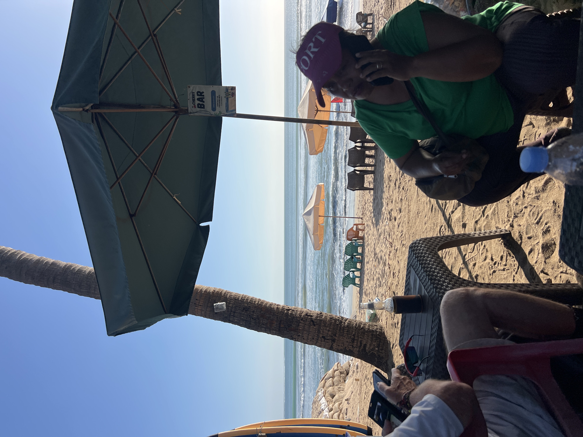 The image depicts a beach scene in Kuta, Indonesia, on a sunny day. In the foreground, a person wearing a green t-shirt and a purple cap is sitting and talking on a phone. To their left, another person, partially visible, is seated and looking at a mobile phone. A small plastic table with a bottle of beer and a phone charging cable is between them. In the background, the ocean stretches to the horizon under a clear blue sky. Several beach chairs and umbrellas are set up on the sand, with a palm tree to the left of the frame. A surfboard stands upright near the palm tree. A sign for "CHERRY BAR" is visible under a large umbrella. The sand appears to be light brown and there are some sandbags and a light-colored surfboard stacked near the water's edge.