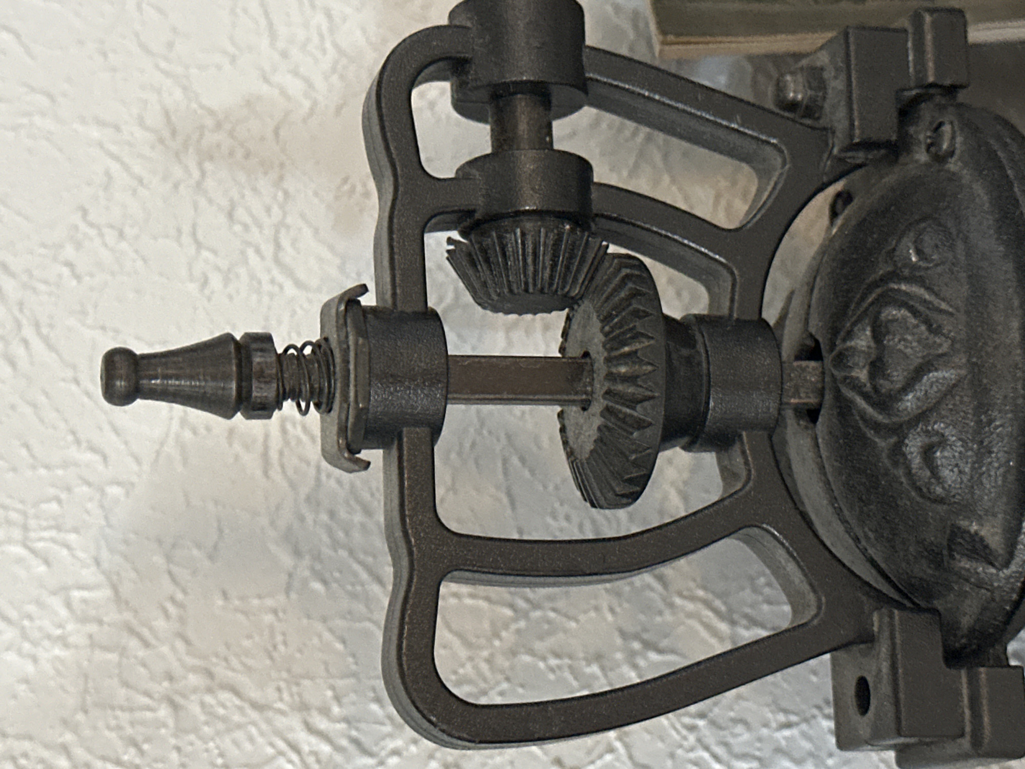 A close-up, head-on, eye-level shot shows the top mechanism of an antique cast-iron coffee grinder. The grinder is dark gray and weathered, with visible signs of age. The background is a textured, off-white wall. The main focus is on the gears and the central shaft of the grinder. A small coil spring is visible around the central shaft, just below a metal bracket. Two beveled gears are prominently displayed. One is attached to the central shaft, and the other is engaged with a horizontal rod extending to the right. The top of the central shaft is capped with a decorative, knob-like finial. The base of the grinder, partially visible at the bottom, has an embossed heart design. The overall impression is one of a well-worn, functional antique. The image does not provide any information about the city of Nevinnomyssk, Russia.