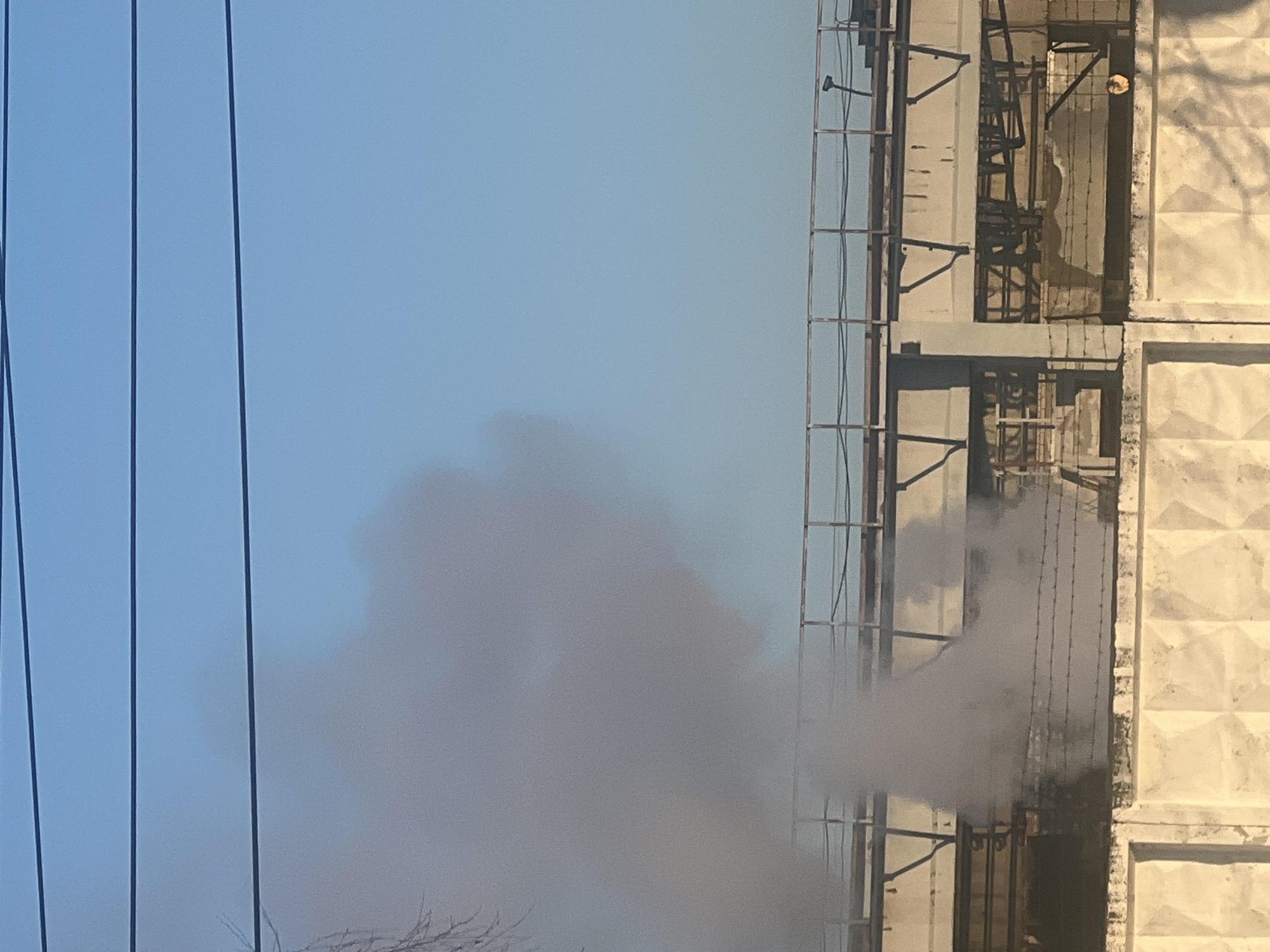 Image depicts an industrial or utility building releasing grey smoke. The building features a concrete facade with a grid-like pattern and metal framework, set against a clear blue sky. Electrical wires are visible on the left. The image does not contain people, actions, or interactions. The geographical location in Chaikovsky, Russia, is not directly identifiable from the image.