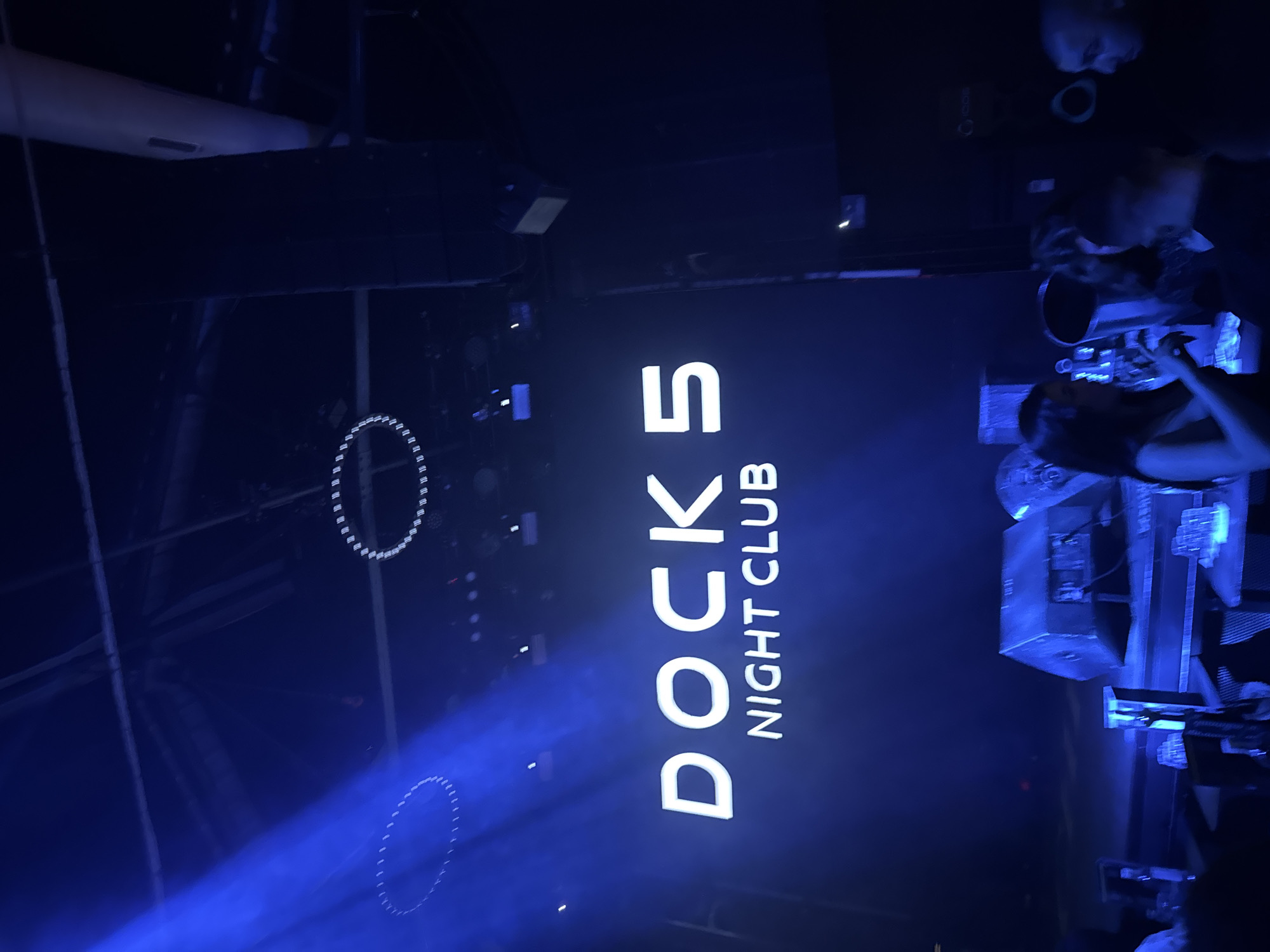 The image depicts the interior of "Docks 5 Night Club," a venue located in Burgas, Bulgaria. The camera captures a view of the club's main screen, which displays the club's name in large, illuminated white letters: "DOCKS 5 NIGHT CLUB." Two large, circular lighting fixtures, constructed with what appear to be individual LED lights, are visible. The lighting in the club is predominantly blue. There are people present, particularly on the right side of the frame, where it looks like a performance or DJ set is taking place. Electronic equipment, likely part of the sound and lighting setup, is also visible on the right.